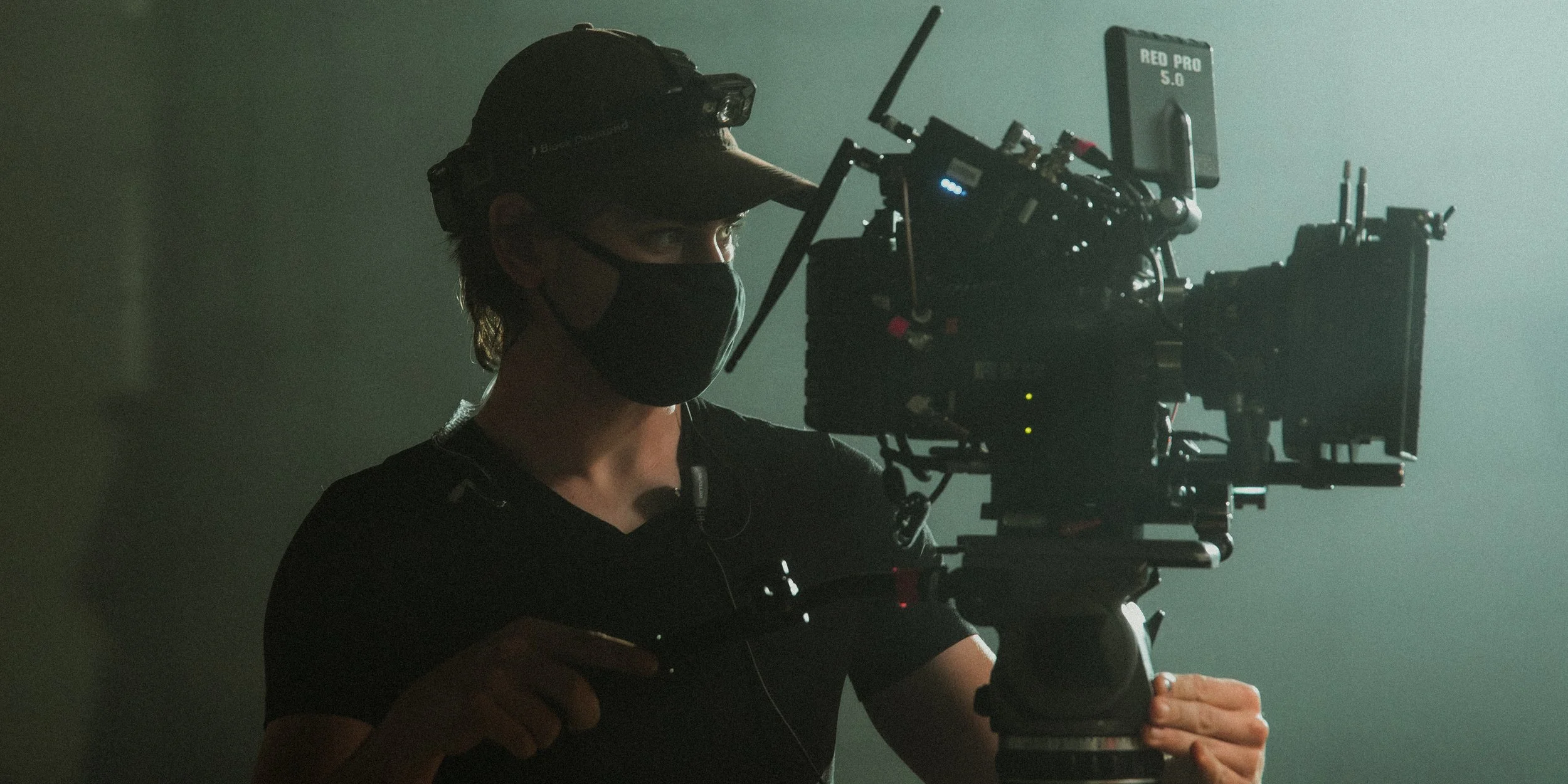 A person wearing a black face mask, cap, and old-style headphones operating a professional camera on a dark set.