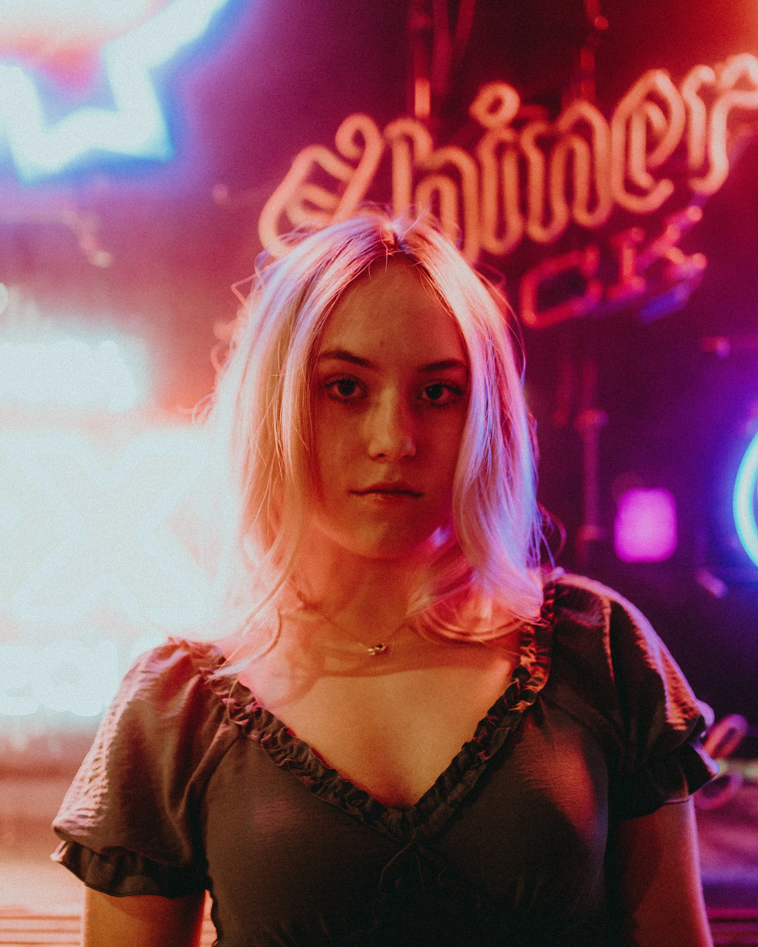 A woman with blonde hair looking directly at the camera, standing in front of colorful neon lights with a partially visible sign in the background.