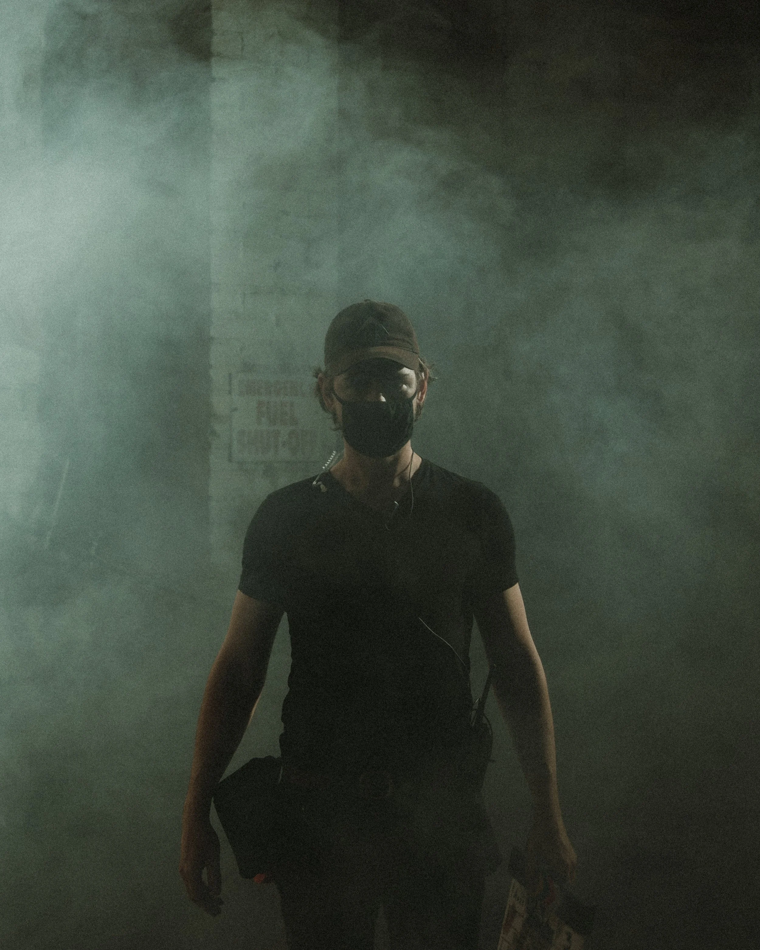 A person wearing a black face mask, a dark baseball cap, and black shirt standing in a foggy or smoky environment.