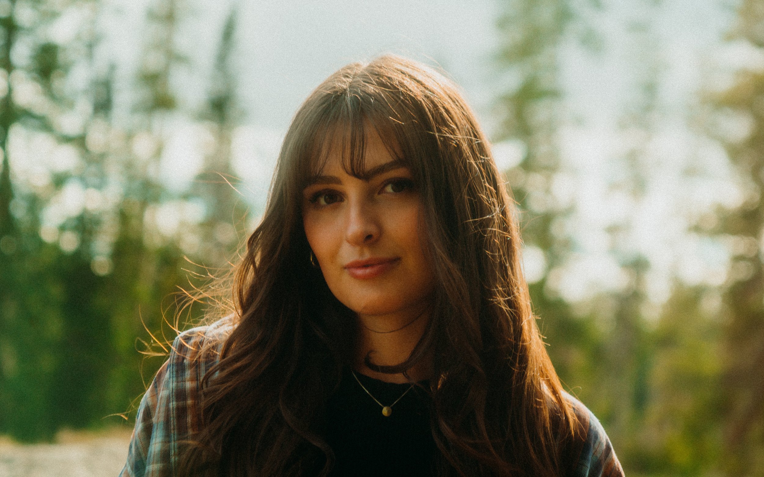 A young woman with long brown hair and bangs, wearing a plaid shirt and necklace, outdoors with sunlight filtering through trees behind her.