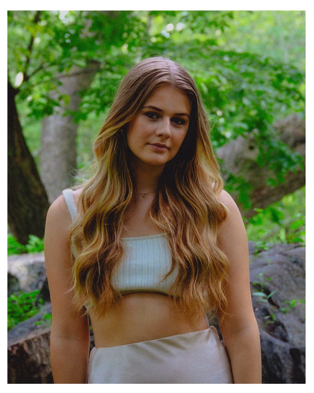 A young woman with long, wavy, blonde hair, wearing a white tank top, standing outdoors with green foliage and trees in the background.