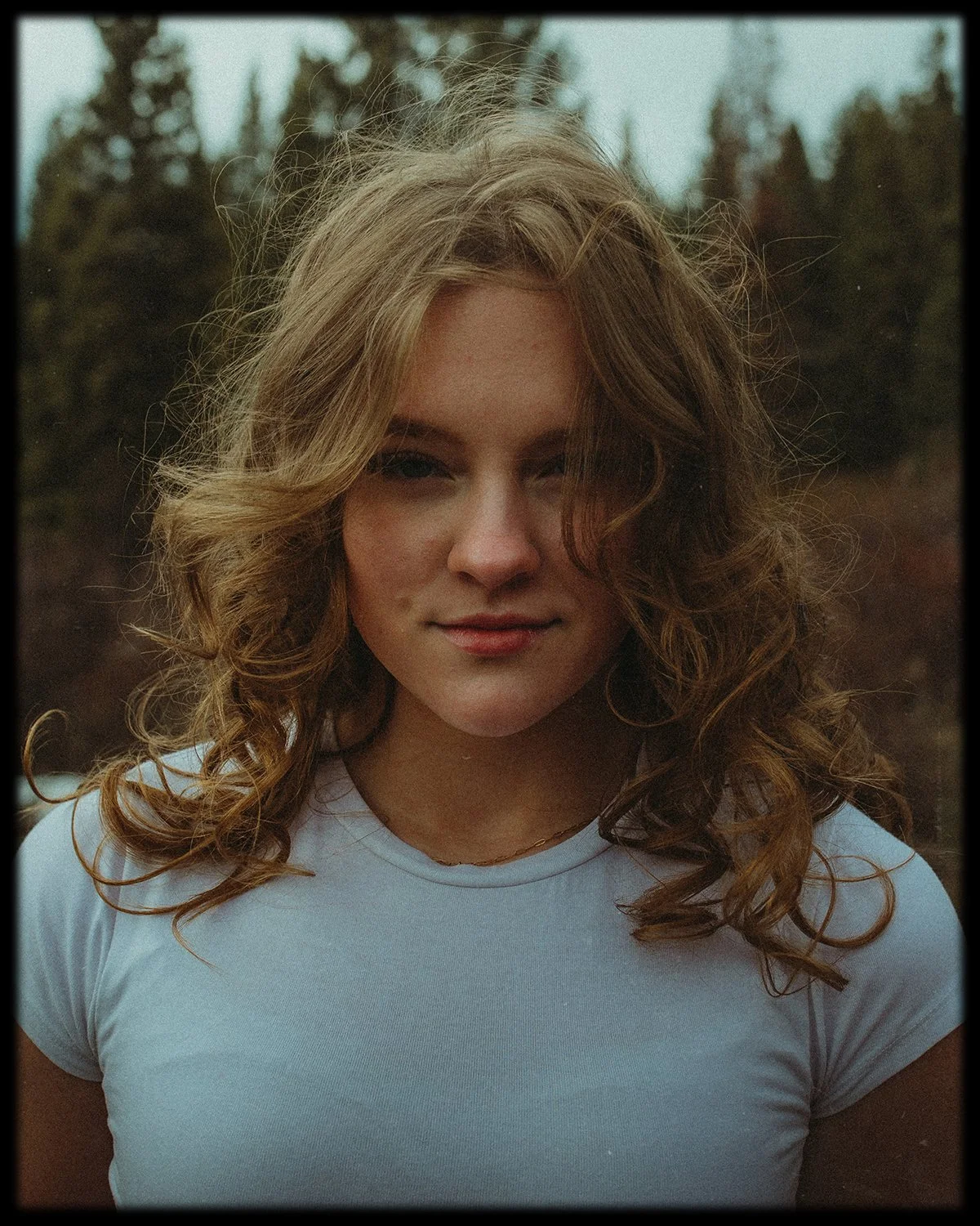 A woman with curly, light brown hair standing outdoors with a blurred forest background. She is wearing a white t-shirt and a gold necklace, with one eye slightly closed and a faint smile.