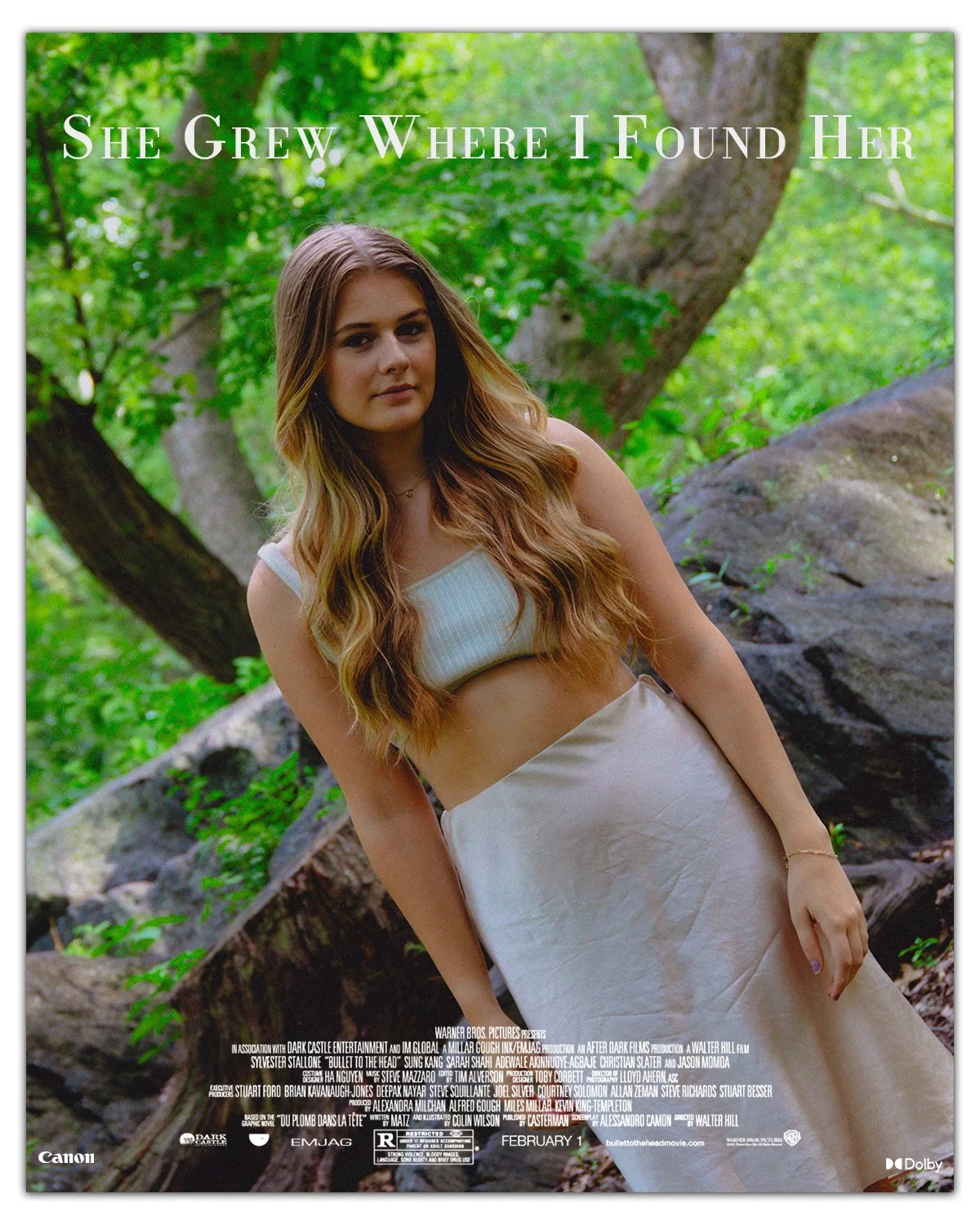 A young woman with long, wavy, light brown hair standing outdoors in a forested area, wearing a white sleeveless crop top and a white skirt, with green foliage and rocks in the background.
