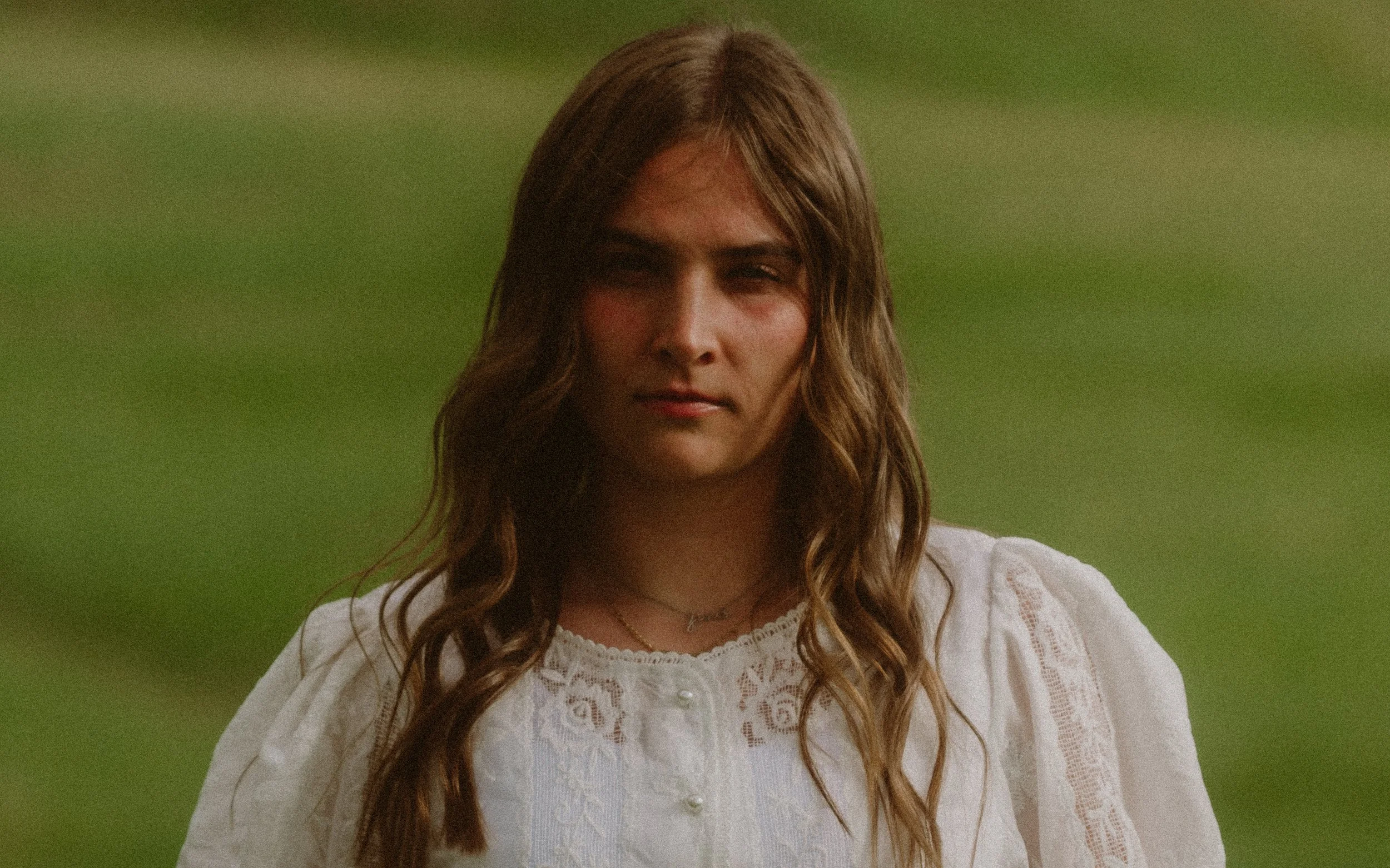 A woman with long wavy hair wearing a white lace top standing outdoors in front of a blurred green background.