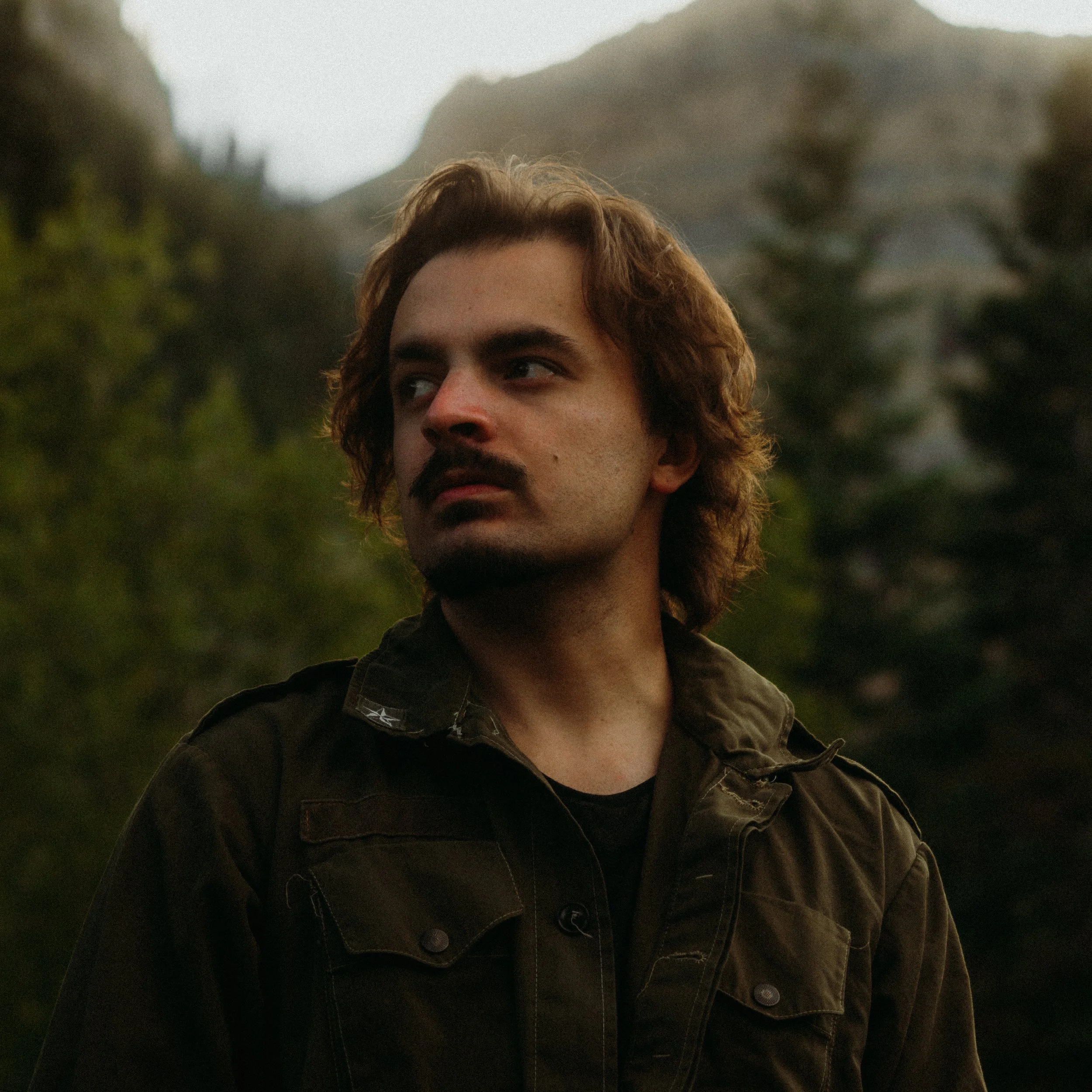Partial view of a young man with brown, wavy hair and a mustache, wearing a dark jacket, outdoors with blurry trees and mountains in the background.