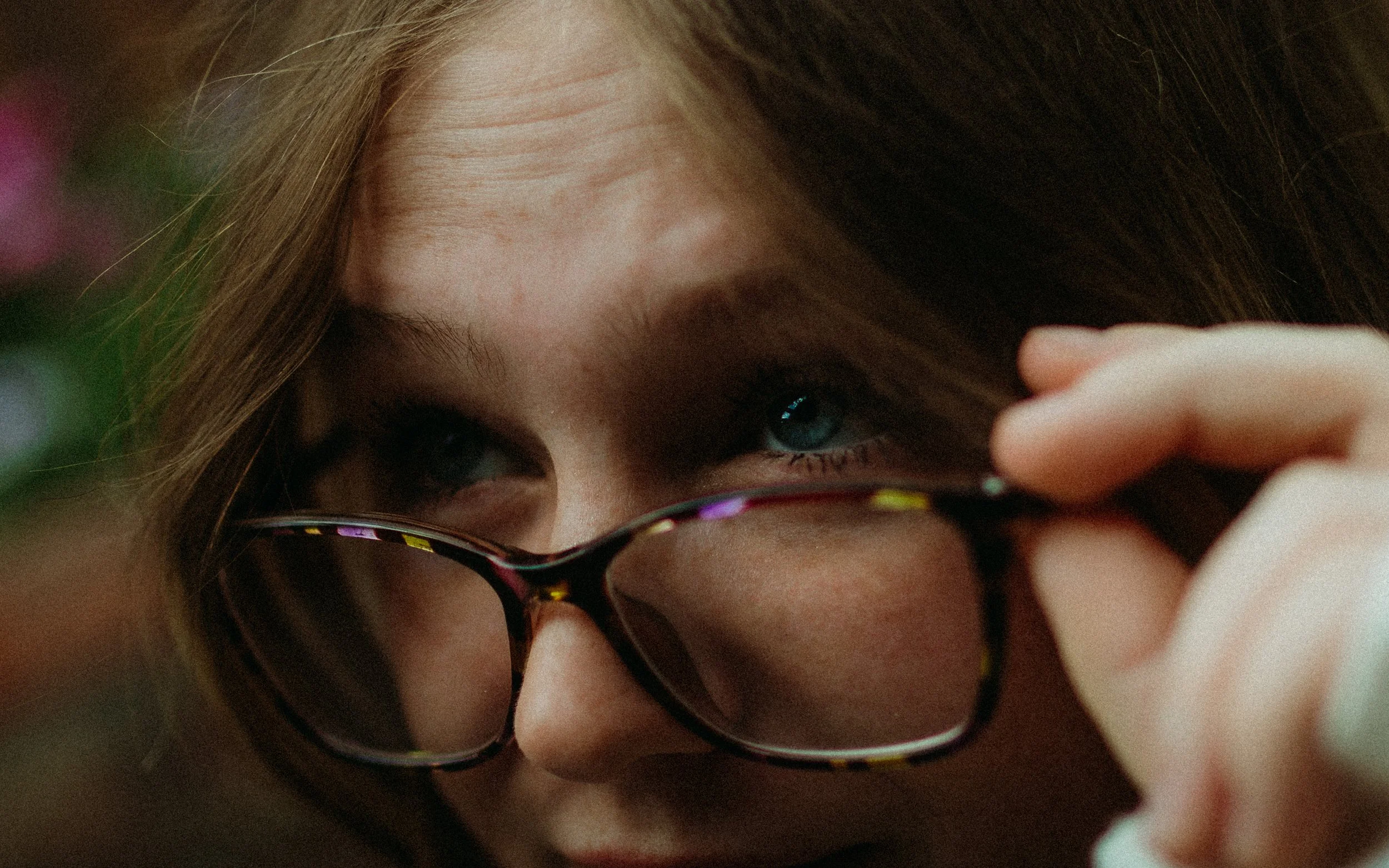 A person with brown hair and blue eyes, wearing with dark glasses, is adjusting or lowering their glasses with their hand.