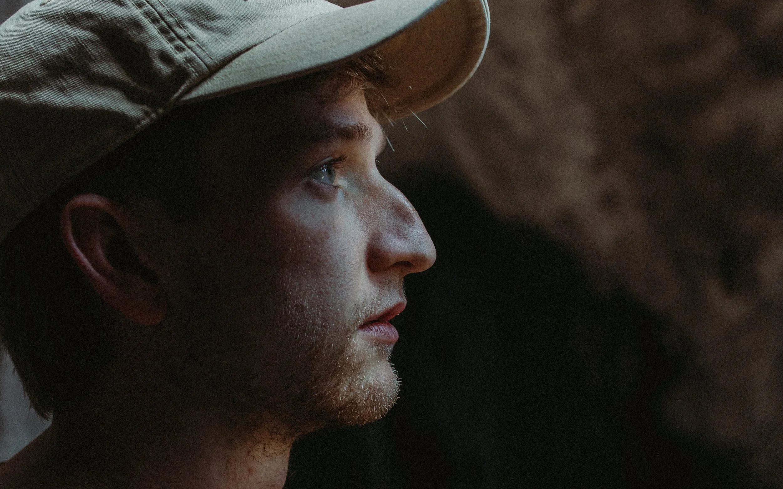 Profile of a young man with fair skin, facial hair, and blue eyes, wearing a tan baseball cap, in a dark setting.