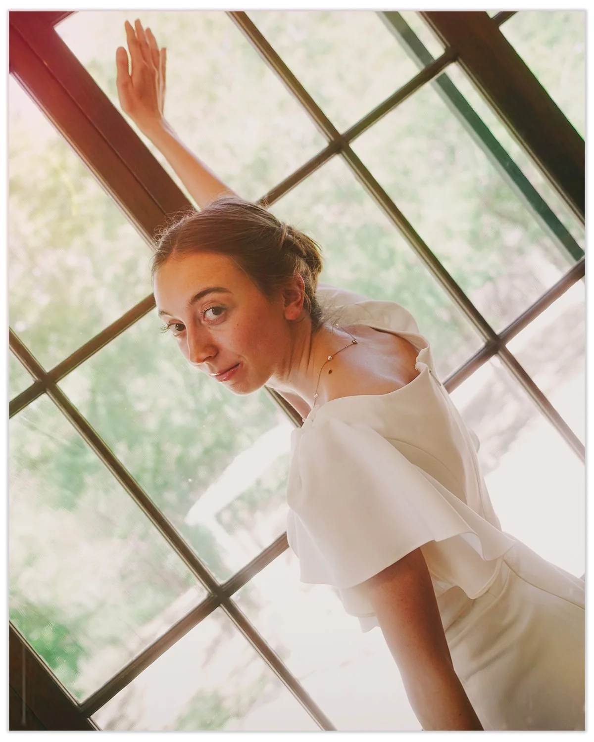 A woman with brown hair styled in an updo, wearing a white off-the-shoulder dress, standing near a large window with multiple panes, with greenery visible outside.