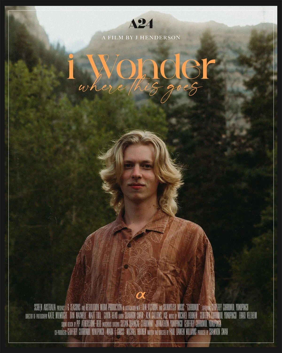 Young man with long blonde hair standing outdoors in a scenic forested mountainous area.