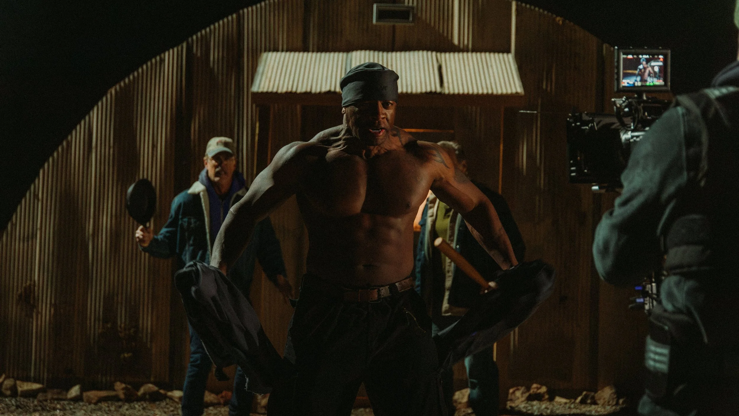 A shirtless man with a black bandana on his head stands in front of a camera on a film set, with a group of people behind him in an indoor wooden room.