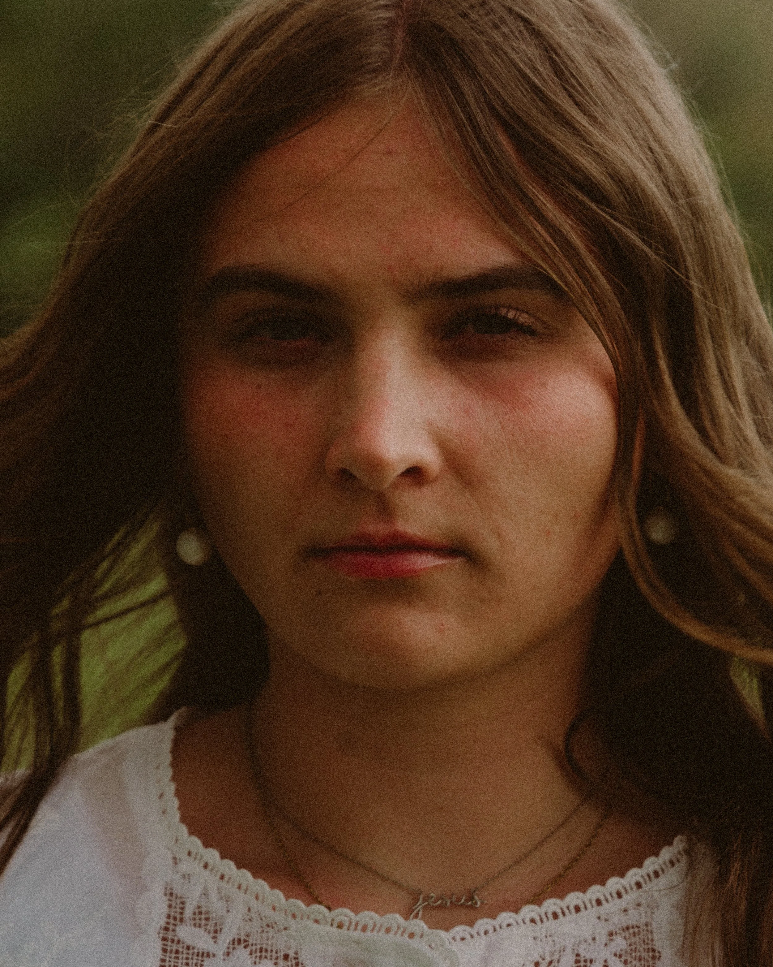 Close-up of a young woman with light brown hair, wearing a white embroidered top and layered necklaces, with a subtle smile.