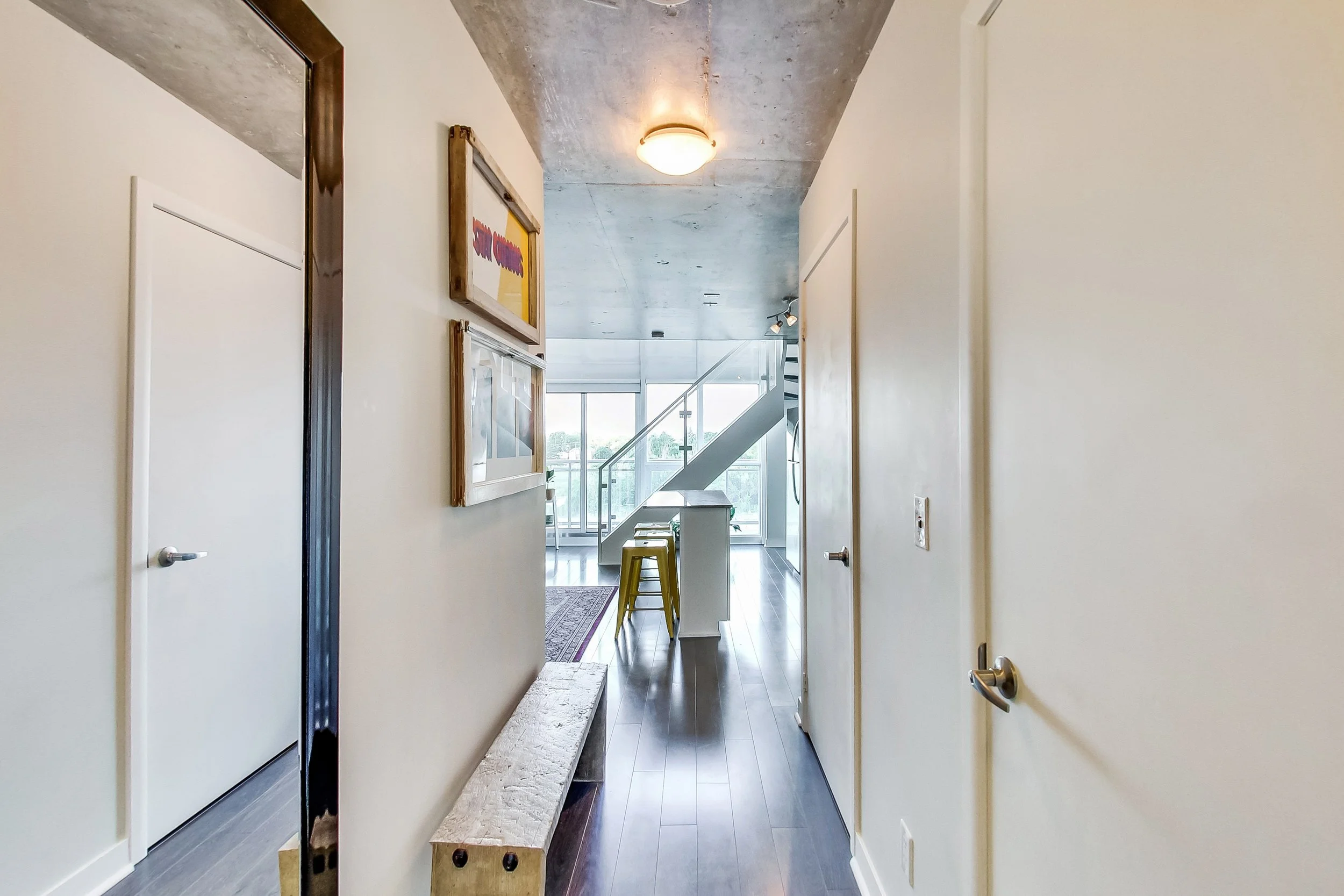 Hallway in the iLOFT Etobicoke renovation with an exposed concrete ceiling, framed art, a glass-railing staircase, and yellow bar stools visible beyond