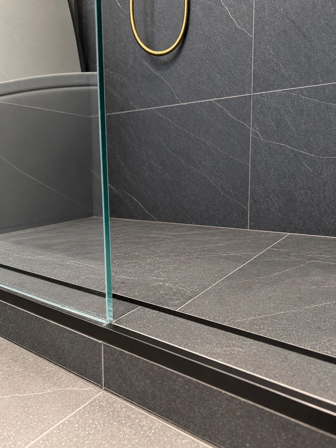 Close-up of a frameless glass shower panel set into a matte black channel on a tiled shower curb, with charcoal stone-look tile and a brushed gold shower hose behind, in the LUX NOIR bathroom renovation by Vogman Design + Build.