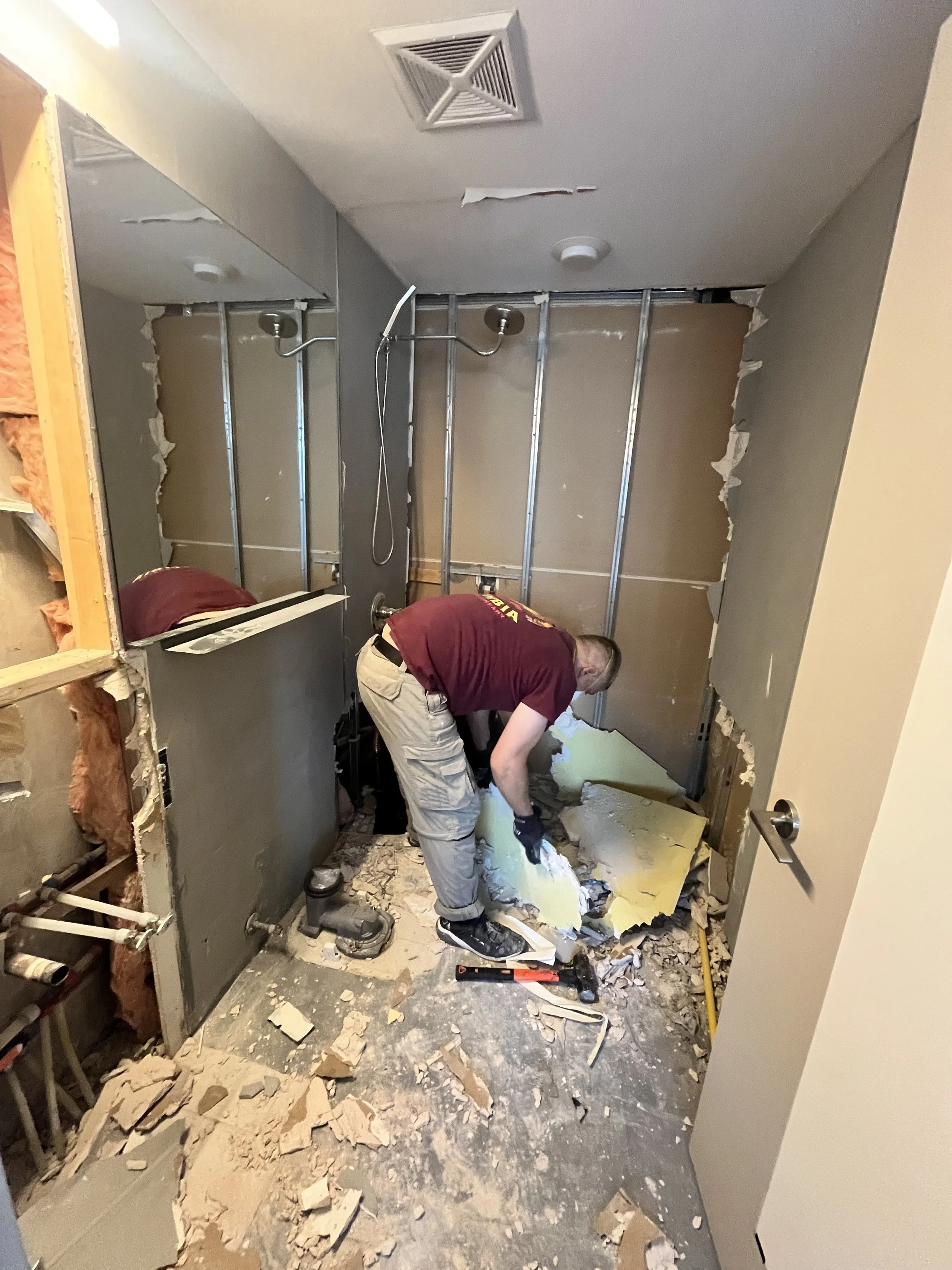 Crew member clearing debris during bathroom demolition, with the bathtub and wall finishes removed to expose the steel studs, plumbing lines and original shower head, during the LUX NOIR bathroom renovation by Vogman Design + Build.