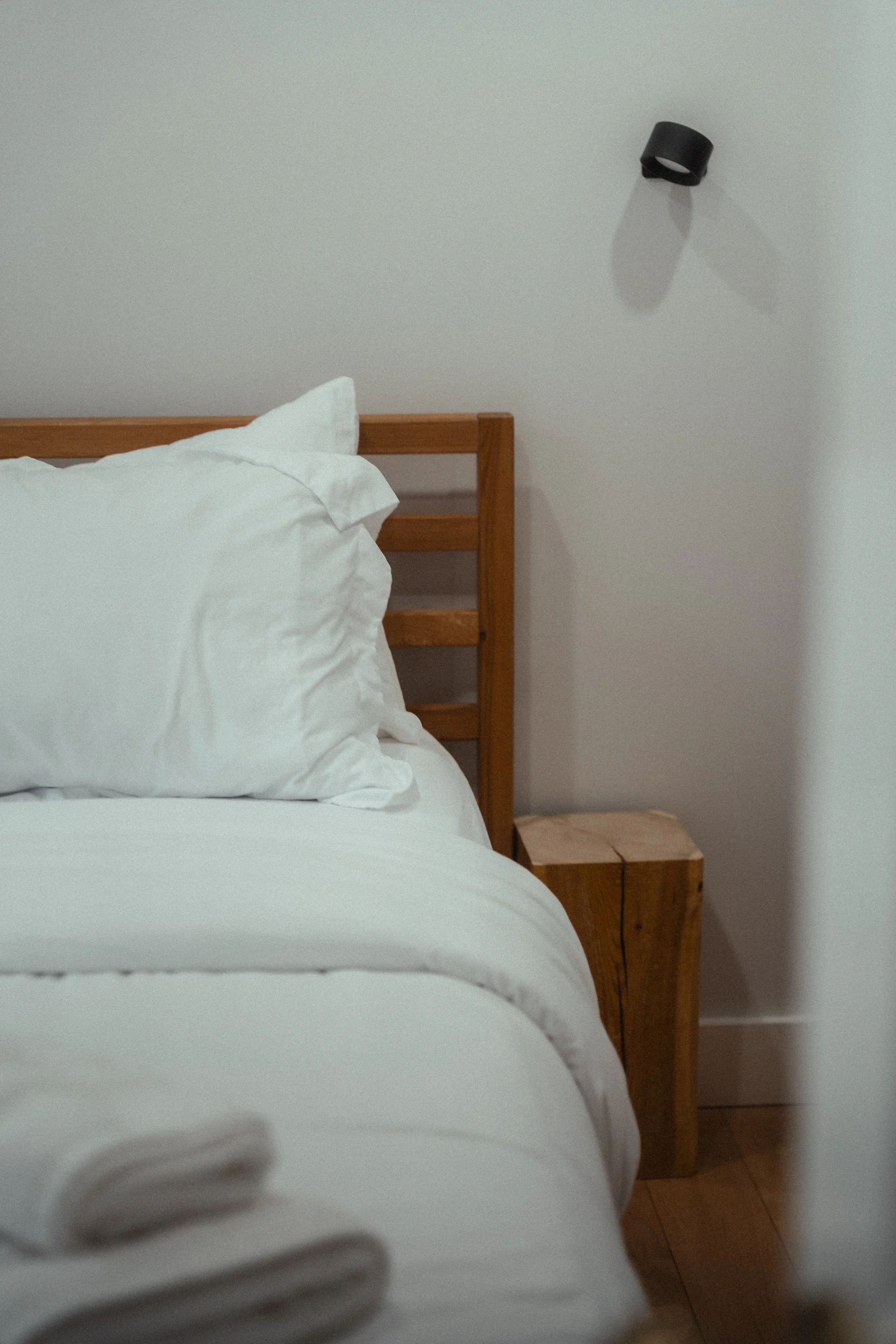 Bedroom detail with white linens, a wood slatted headboard, a live-edge wood nightstand, and a black wall sconce