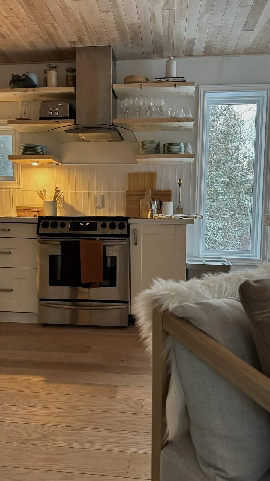 Kitchen in the Tobermory cottage renovation with a stainless steel range and hood, open wood shelving, white shaker cabinets, and a wood plank ceiling - Vogman Design + Build.