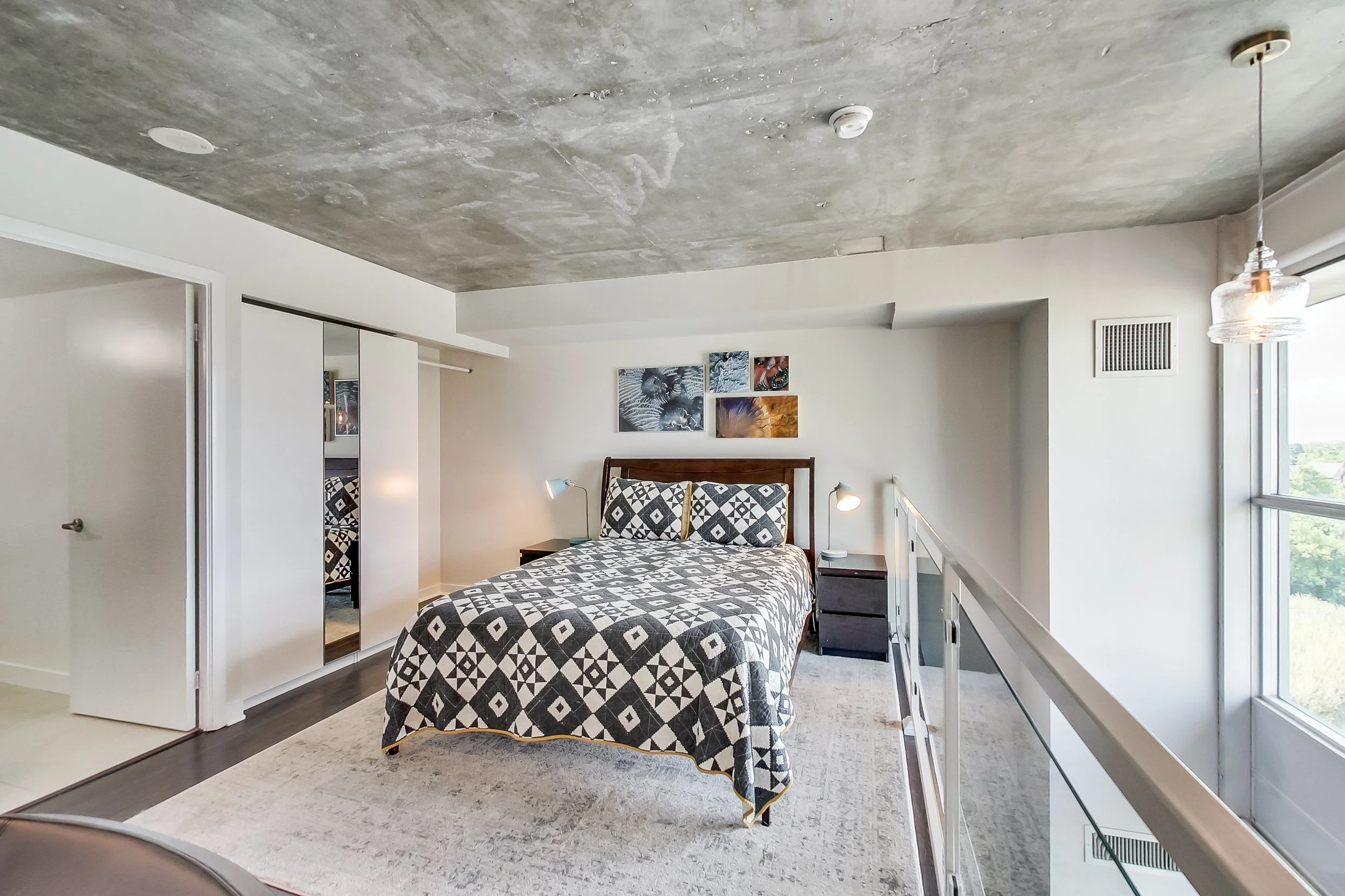 Loft bedroom in the iLOFT Etobicoke renovation with an exposed concrete ceiling, dark wood bed frame, graphic diamond-patterned quilt, and a glass mezzanine railing