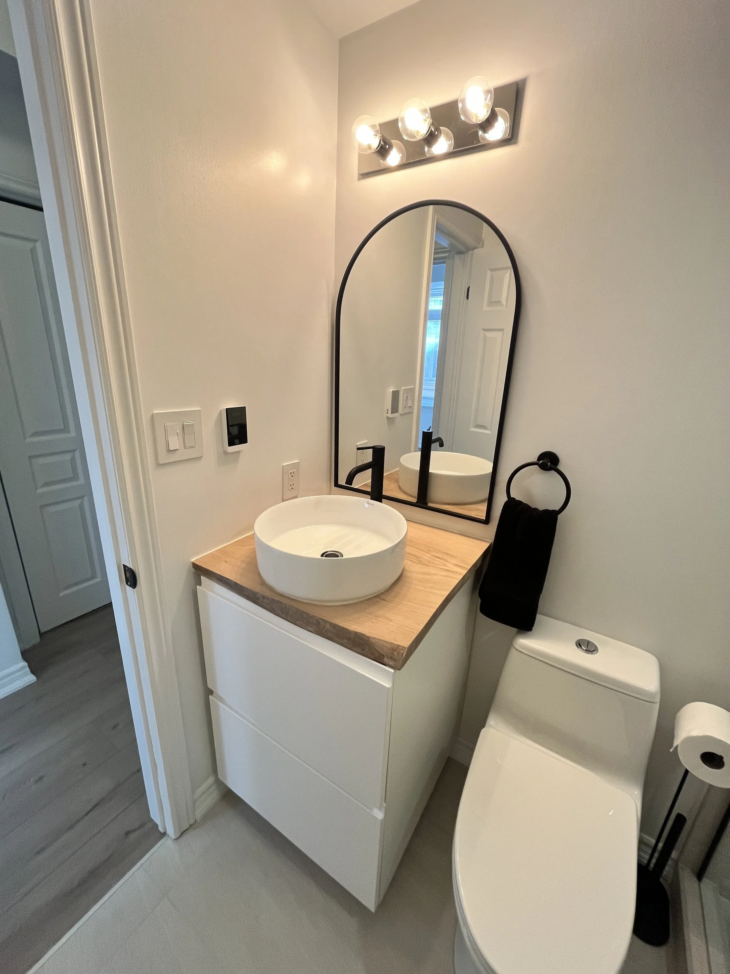 Full view of a Queen West condo bathroom renovation with a floating white vanity, round vessel sink, arched black-framed mirror, and modern toilet
