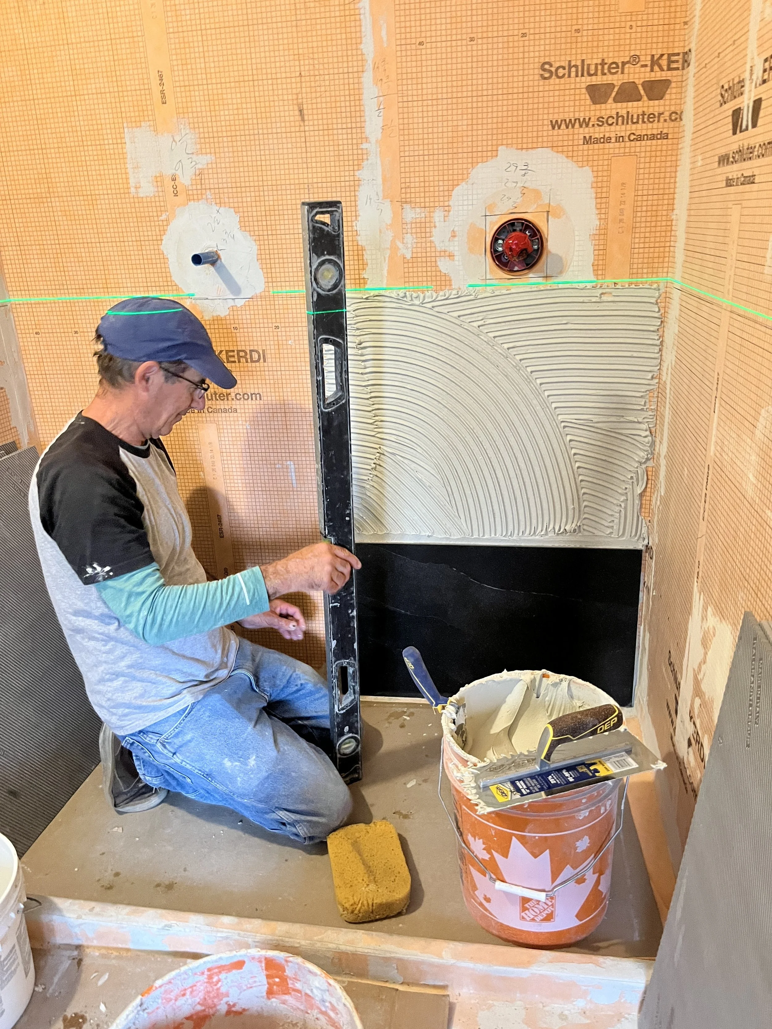 Petre using a level to check wall prep before tiling, with troweled thin-set and Schluter-Kerdi waterproofing membrane visible in a shower installation