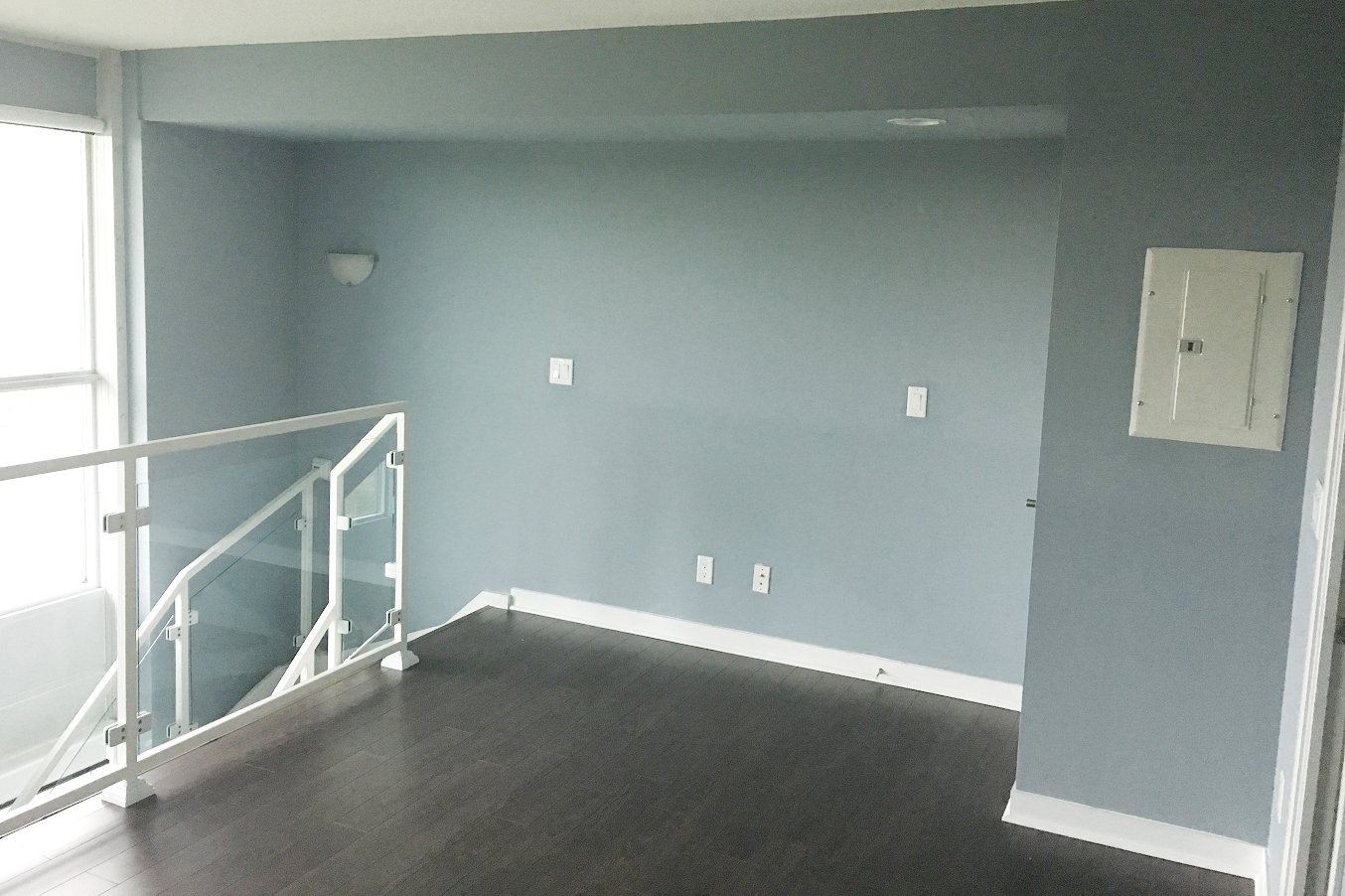 Before photo of an empty loft bedroom nook with blue walls, dark wood flooring, and a glass mezzanine railing, ahead of the full renovation