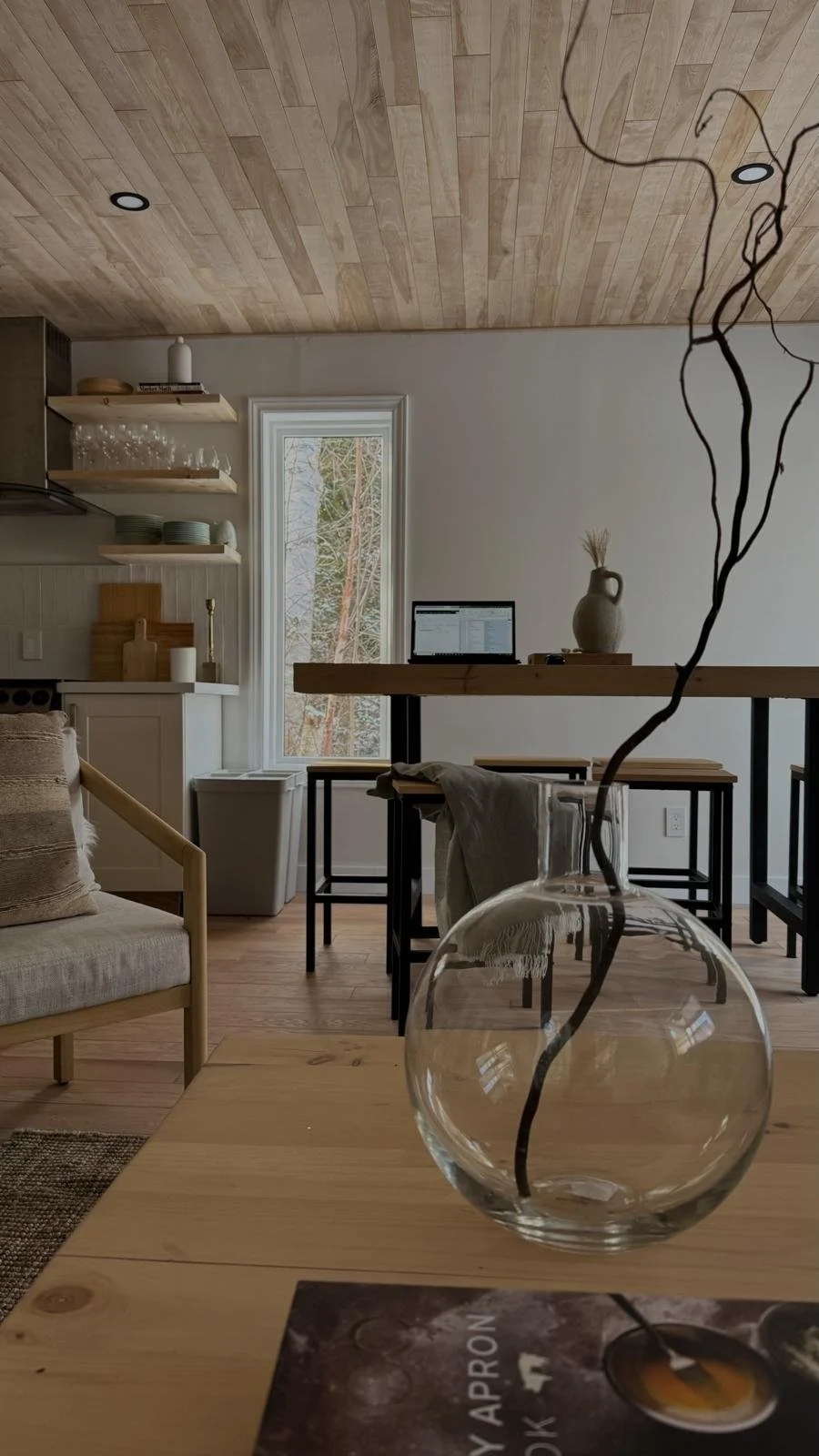 Nordic-inspired living and dining nook in the Tobermory cottage renovation, with a wood plank ceiling, wooden bench dining table, open kitchen shelving, and a glass vase with dried branches - Vogman Design + Build.