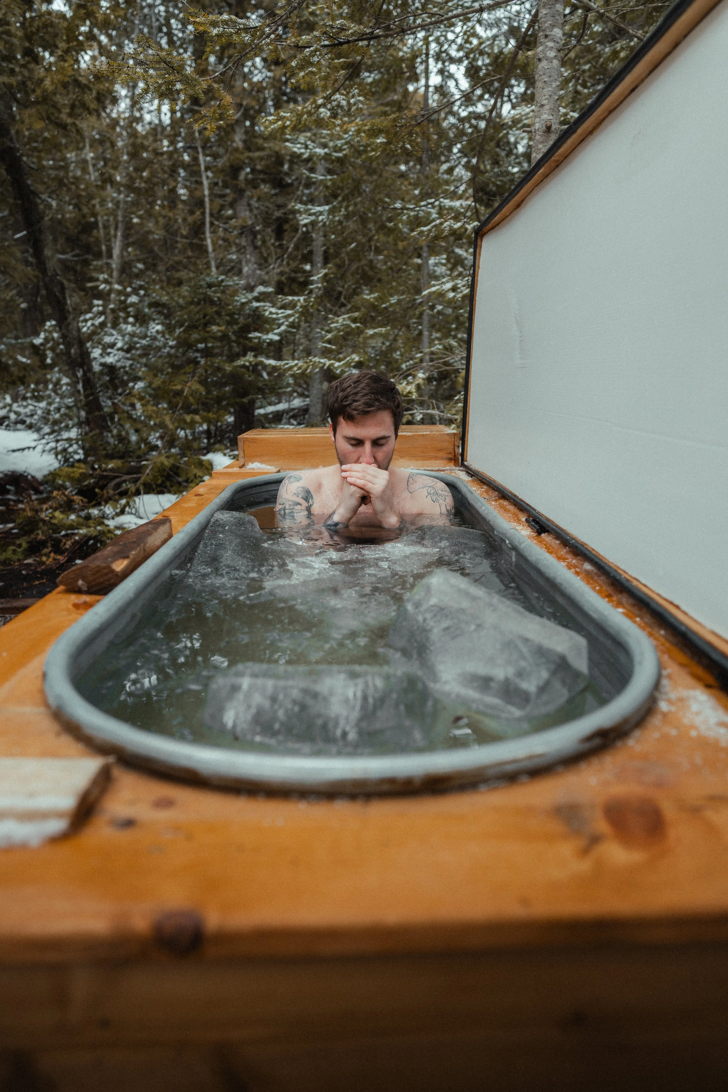 A person sitting in an outdoor cold plunge tub with ice floating on the surface, surrounded by snow-covered trees at the Tobermory cottage renovation