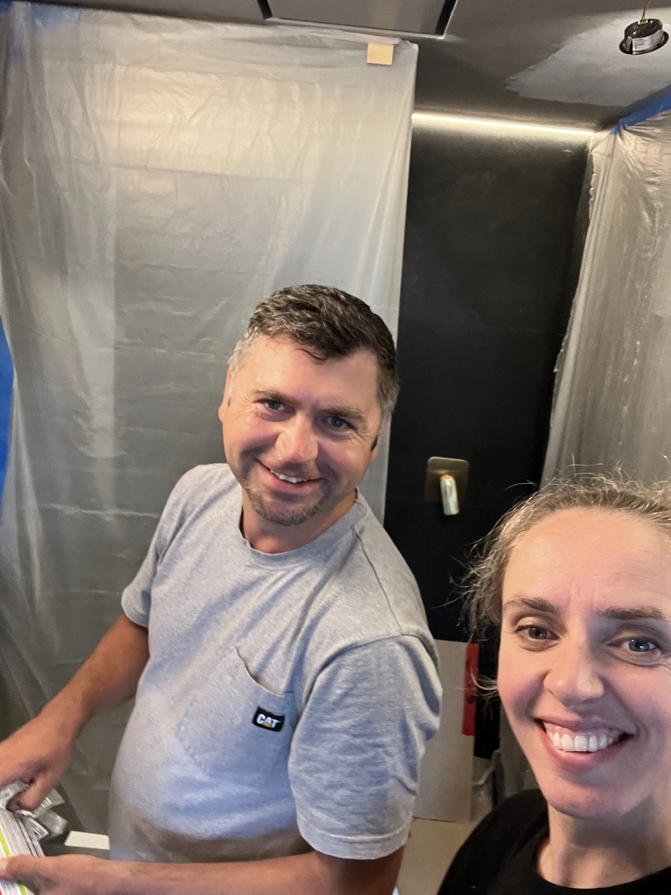 Valentina and Slavik smiling on a job site mid-renovation, with plastic sheeting protecting the space during electrical and lighting work