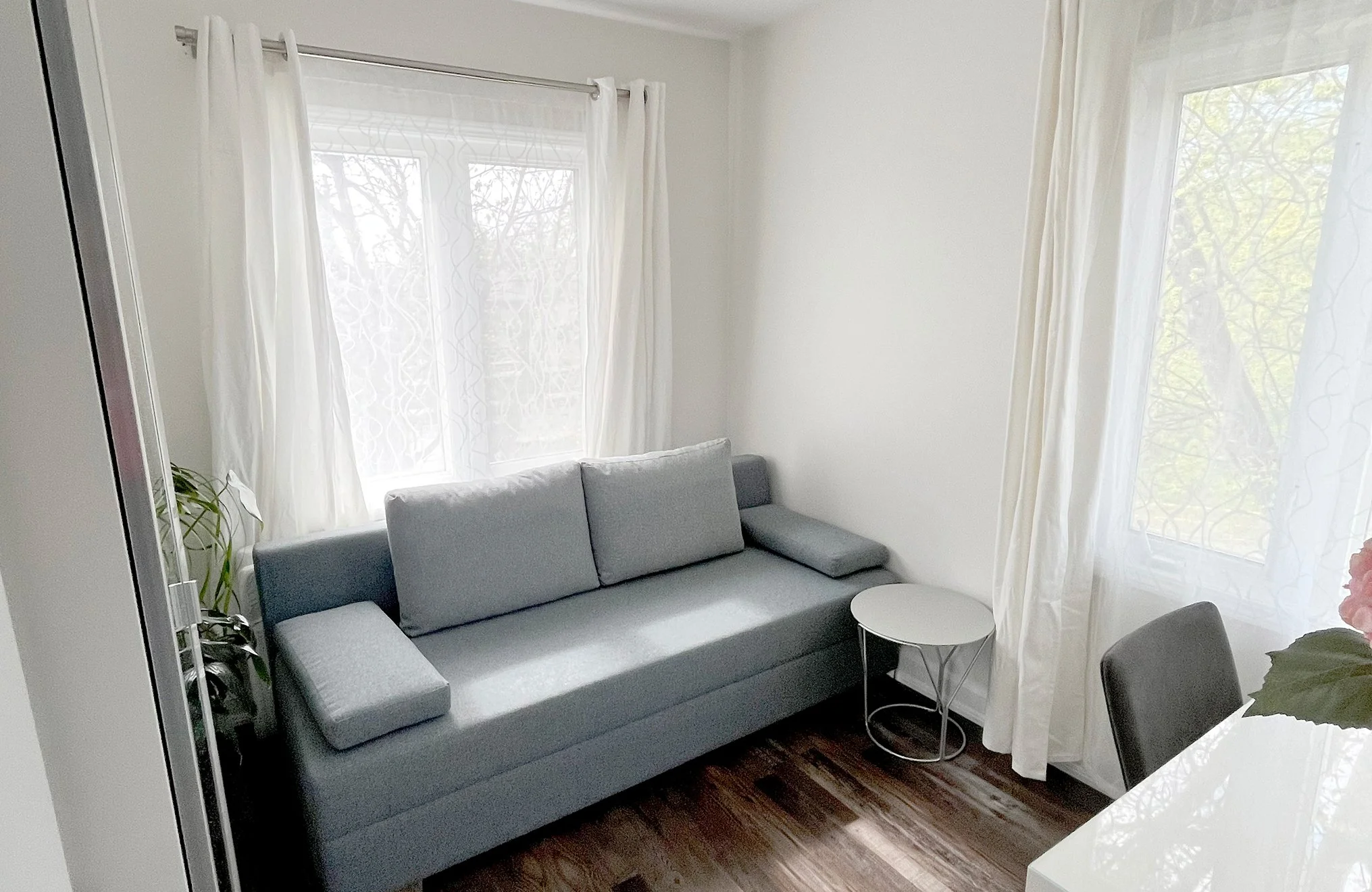 After photo of THE OASIS office renovation with a grey sofa, wood-look flooring, sheer curtains, and a small round side table