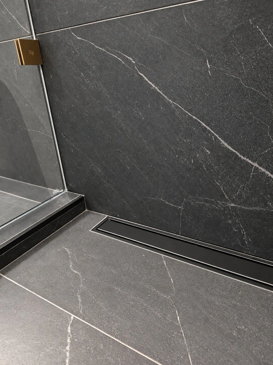 Matte black Schluter linear drain set flush along the shower wall in charcoal stone-look tile with white veining, beside a frameless glass panel with a brushed gold hinge, in the LUX NOIR bathroom renovation by Vogman Design + Build.
