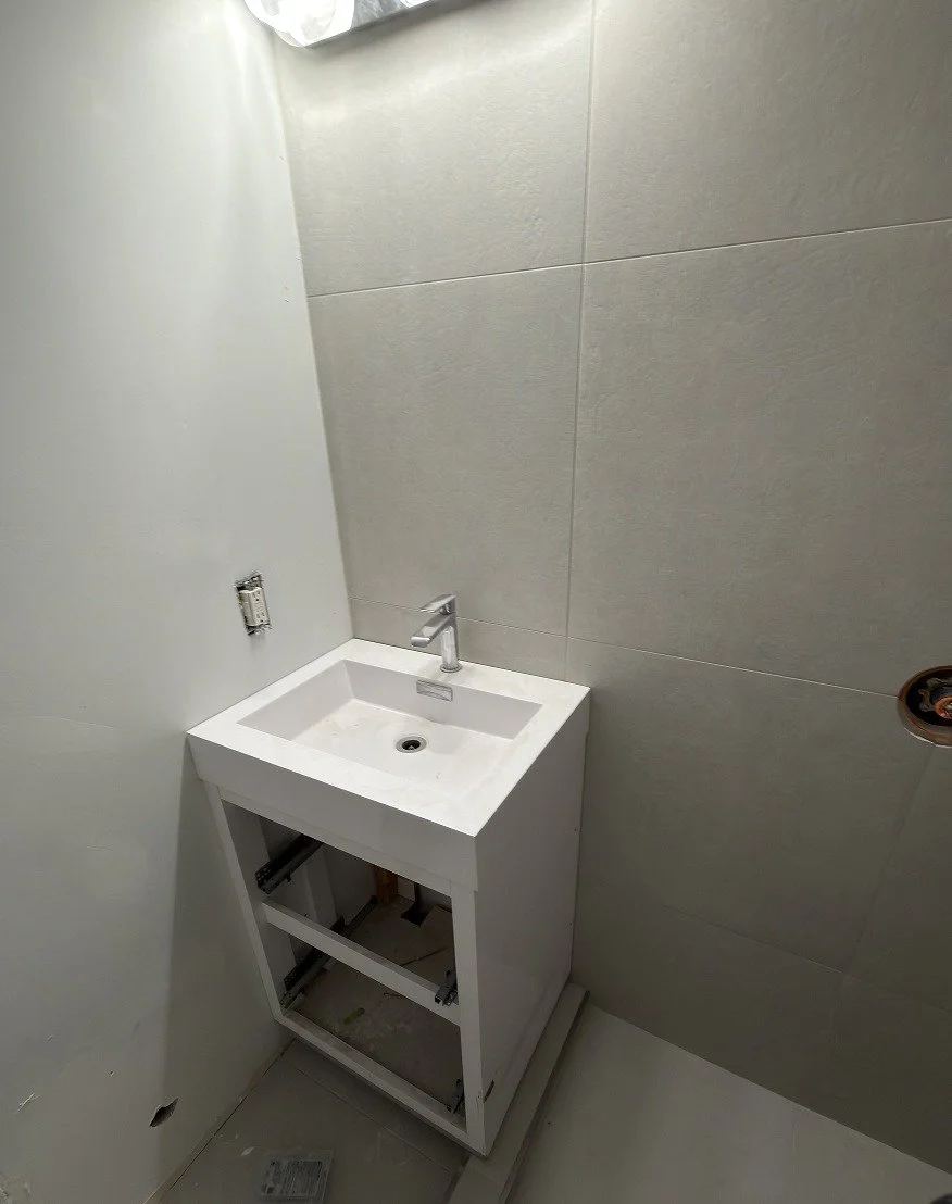 White bathroom vanity with an integrated rectangular sink and chrome single-handle faucet being installed against large-format light grey wall tile, with the drawers not yet fitted, during The Rebuild renovation by Vogman Design + Build.