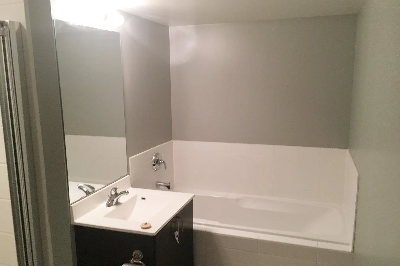 Before photo of the iLOFT bathroom with grey painted walls above white tile, a dark wood vanity, and a built-in bathtub, ahead of the full renovation