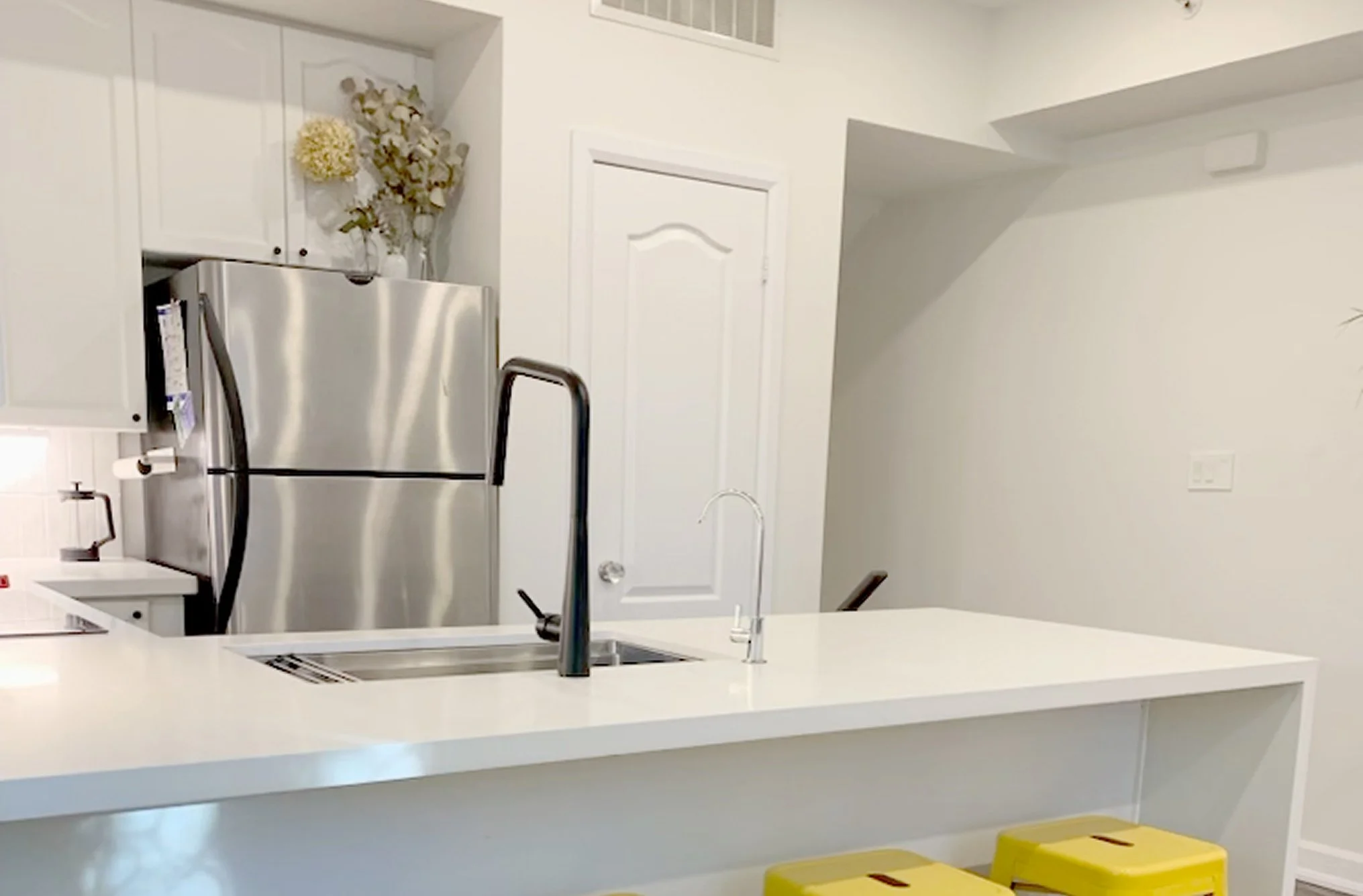 After photo of THE OASIS kitchen renovation with a white waterfall island, matte black and chrome faucets, stainless steel refrigerator, and yellow bar stools