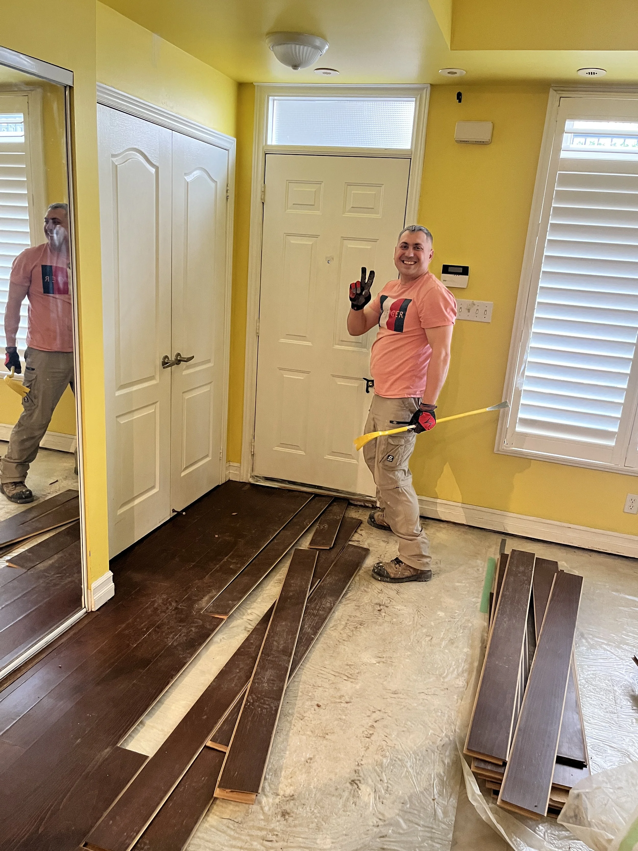 Nikolai smiling and giving a peace sign during a flooring demo, with old laminate planks pulled up and stacked on the floor