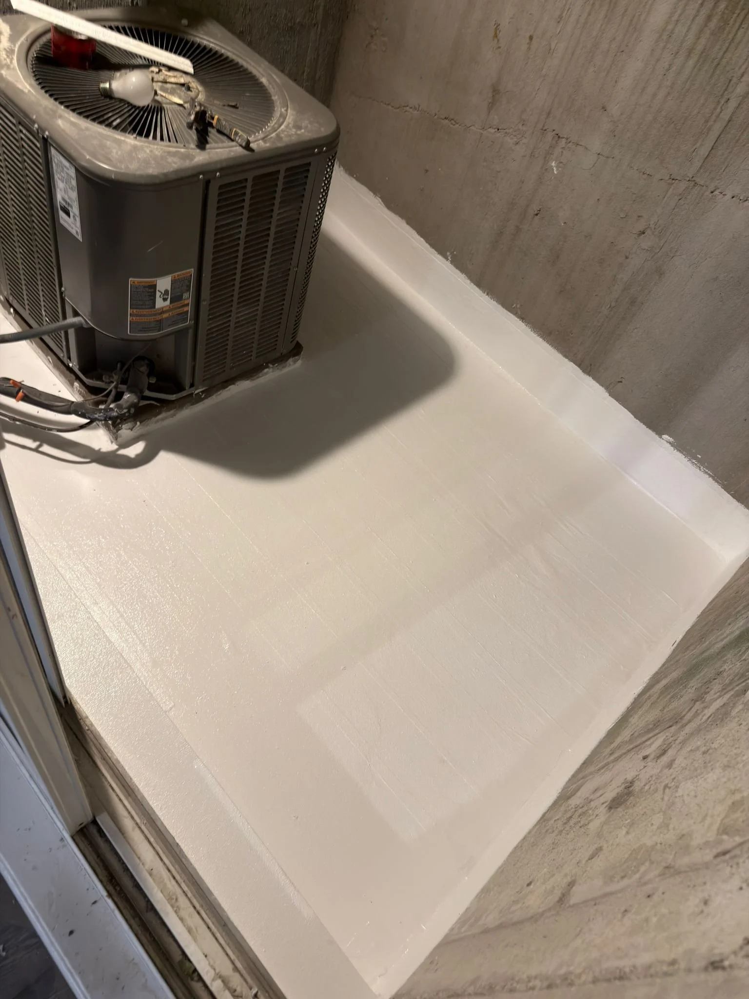 HVAC condenser unit on a freshly waterproofed concrete deck or balcony, with a protective white coating applied around the unit