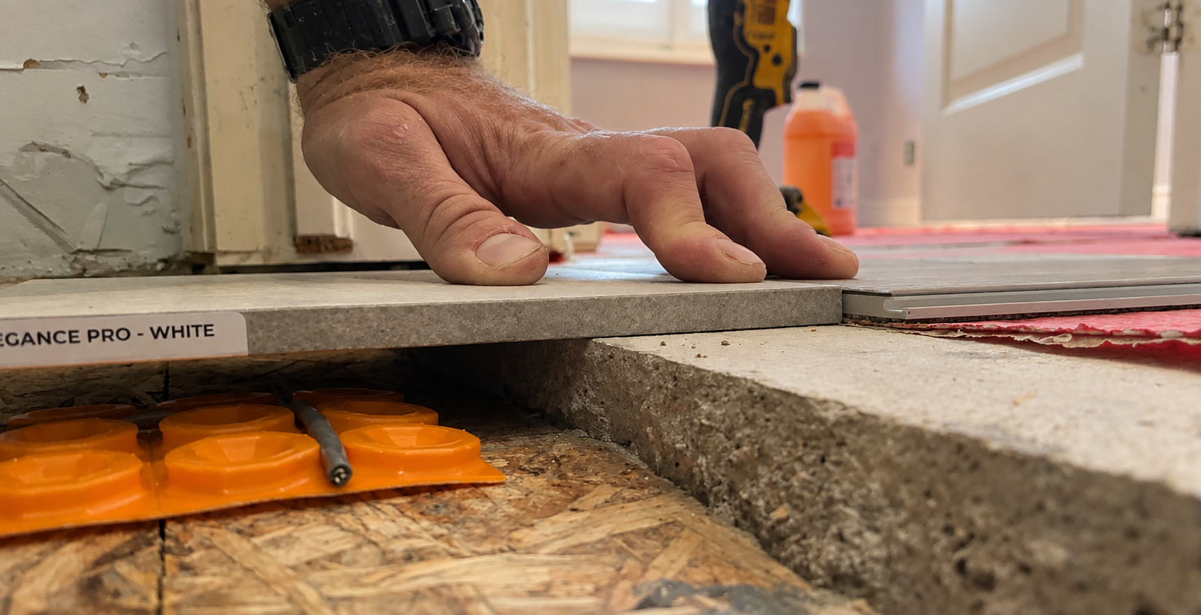 Installer's hand placing a white porcelain tile onto a Schluter uncoupling membrane over the subfloor during the Queen West condo bathroom renovation