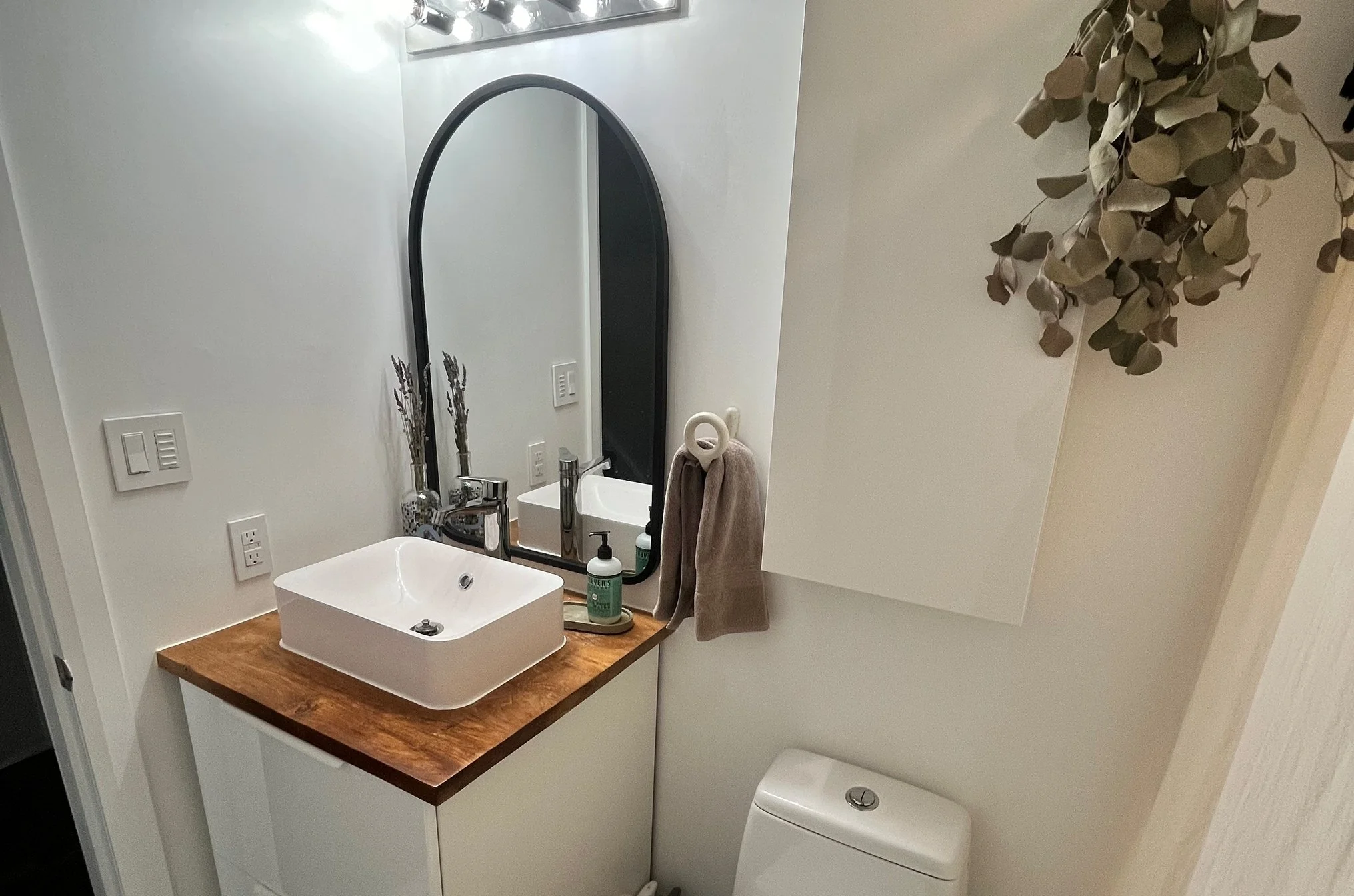 After photo of THE OASIS bathroom renovation with a white vessel sink, wood countertop, arched black-framed mirror, and hanging eucalyptus greenery