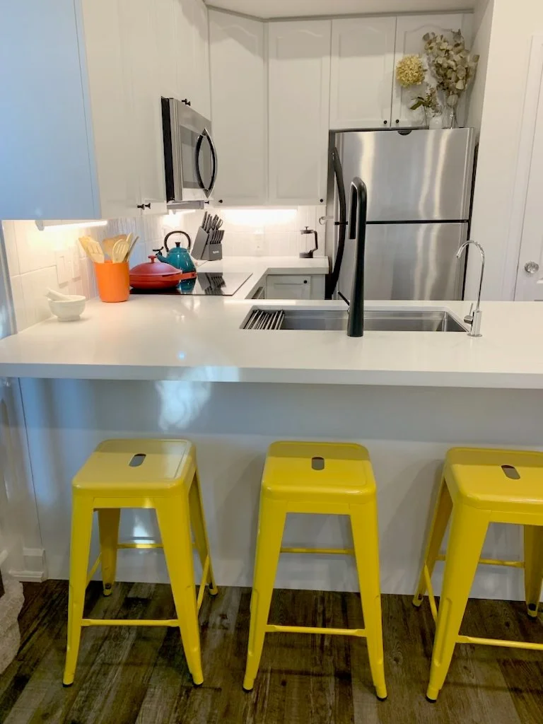 Kitchen in the THE OASIS renovation with white cabinets, stainless steel appliances, a white waterfall countertop, and yellow bar stools