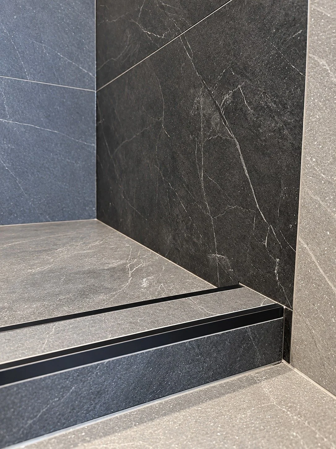 Close-up of a tiled shower curb finished with matte black metal edge trim, where charcoal stone-look shower tile meets lighter grey floor and wall tile, in the LUX NOIR bathroom renovation by Vogman Design + Build.