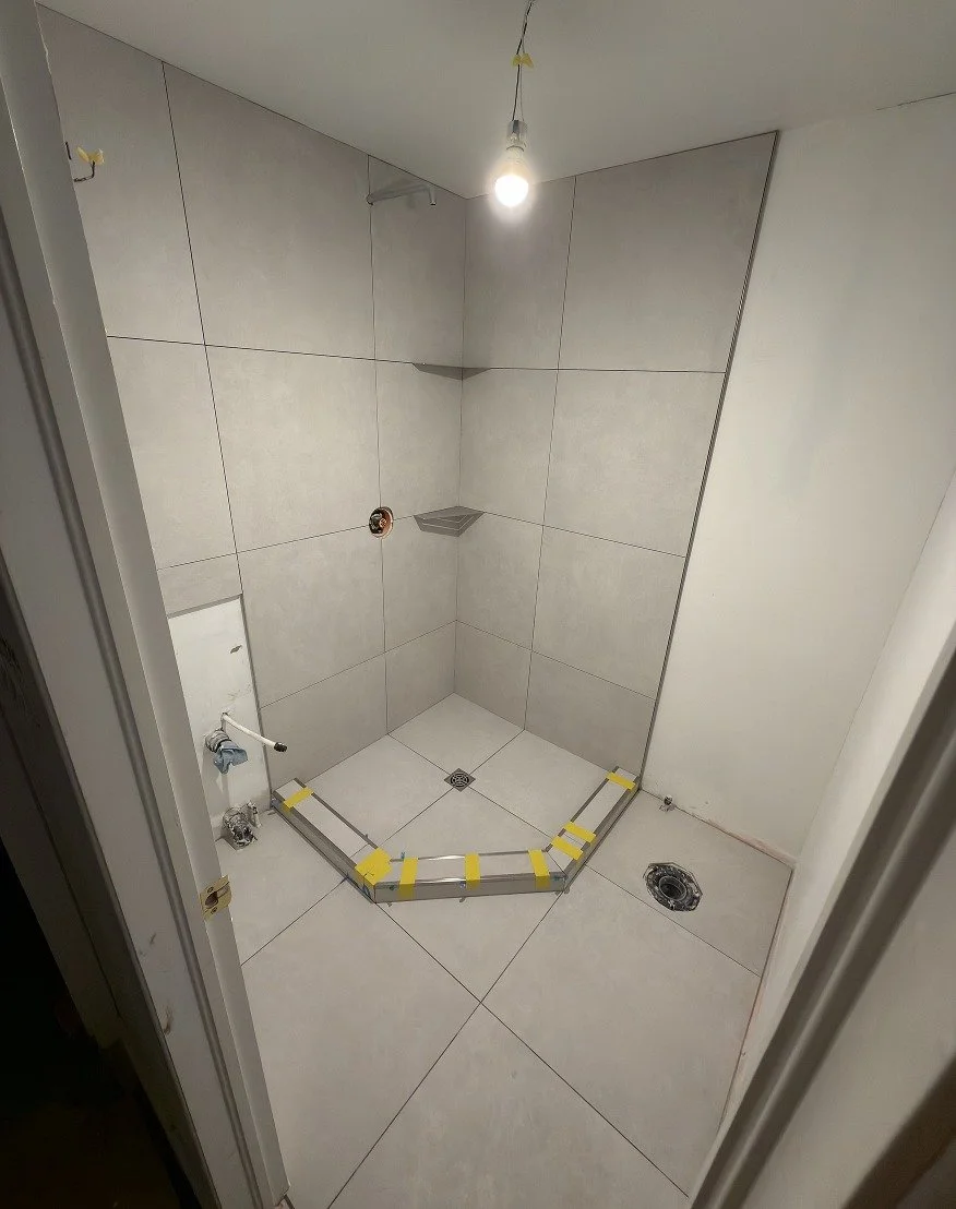 Corner shower tiled in large-format light grey porcelain on the walls and floor, with two corner shelves, a square centre drain and a taped metal curb track in place for the glass enclosure, during The Rebuild renovation by Vogman Design + Build.