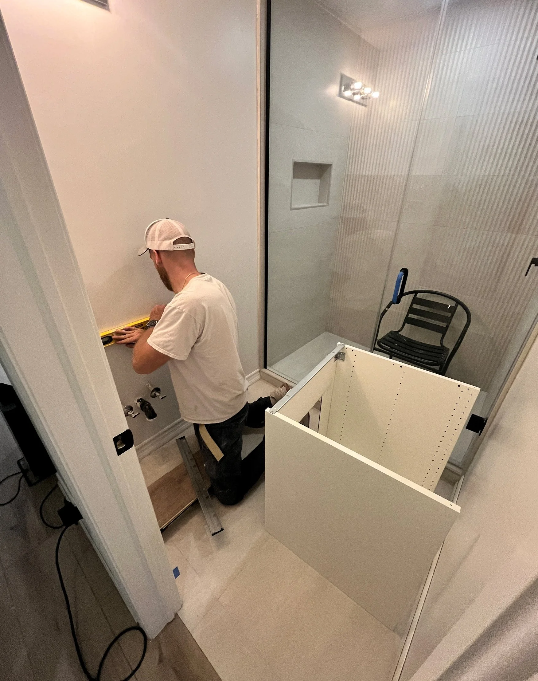 Contractor leveling the wall-mounted plumbing rough-in beside a vanity cabinet frame ready to be installed, with the finished glass shower visible in the background, during the Queen West condo bathroom renovation