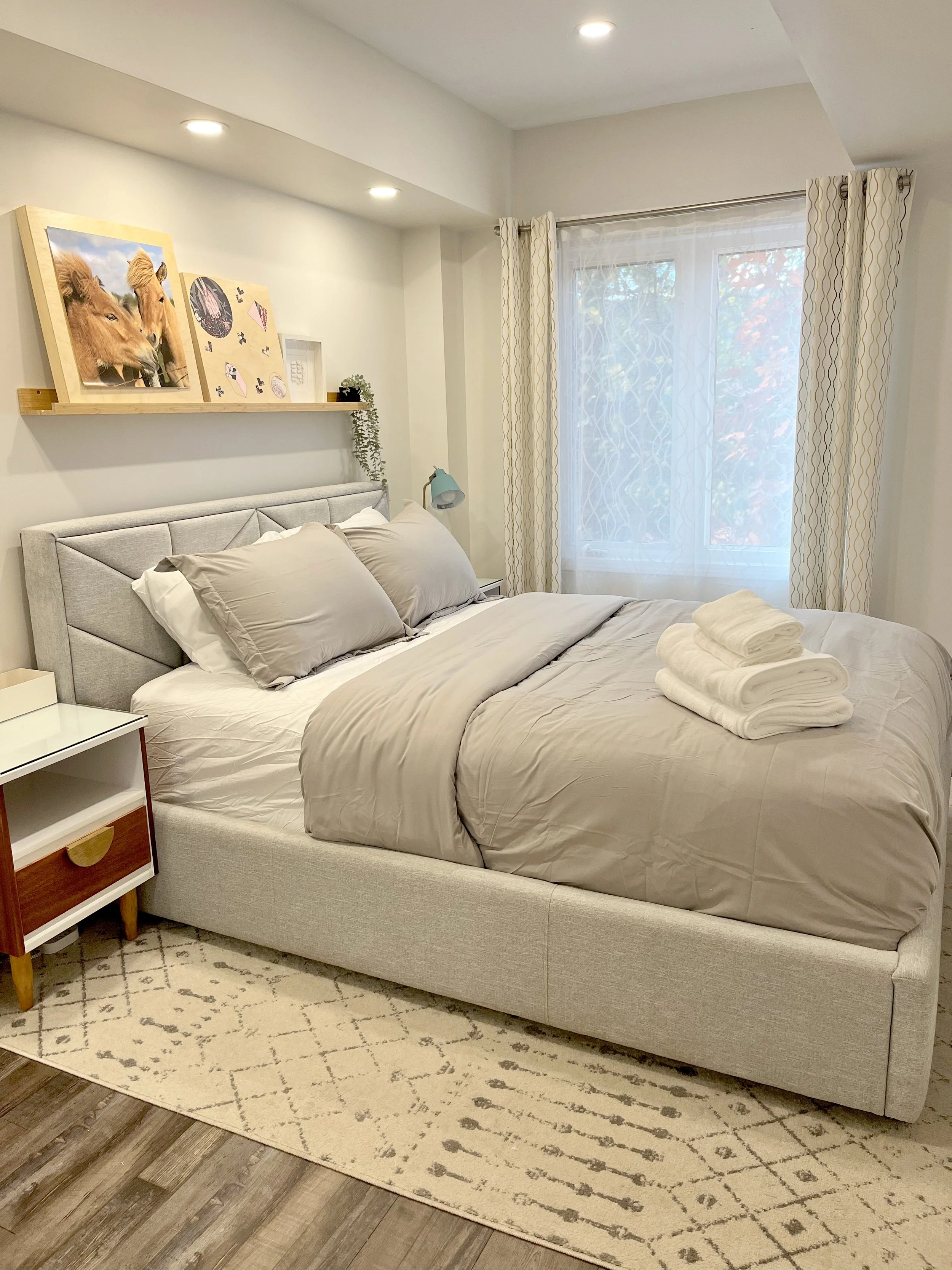 THE OASIS bedroom renovation with a grey upholstered bed, wood floating shelf styled with framed art, a mid-century nightstand, and patterned curtains