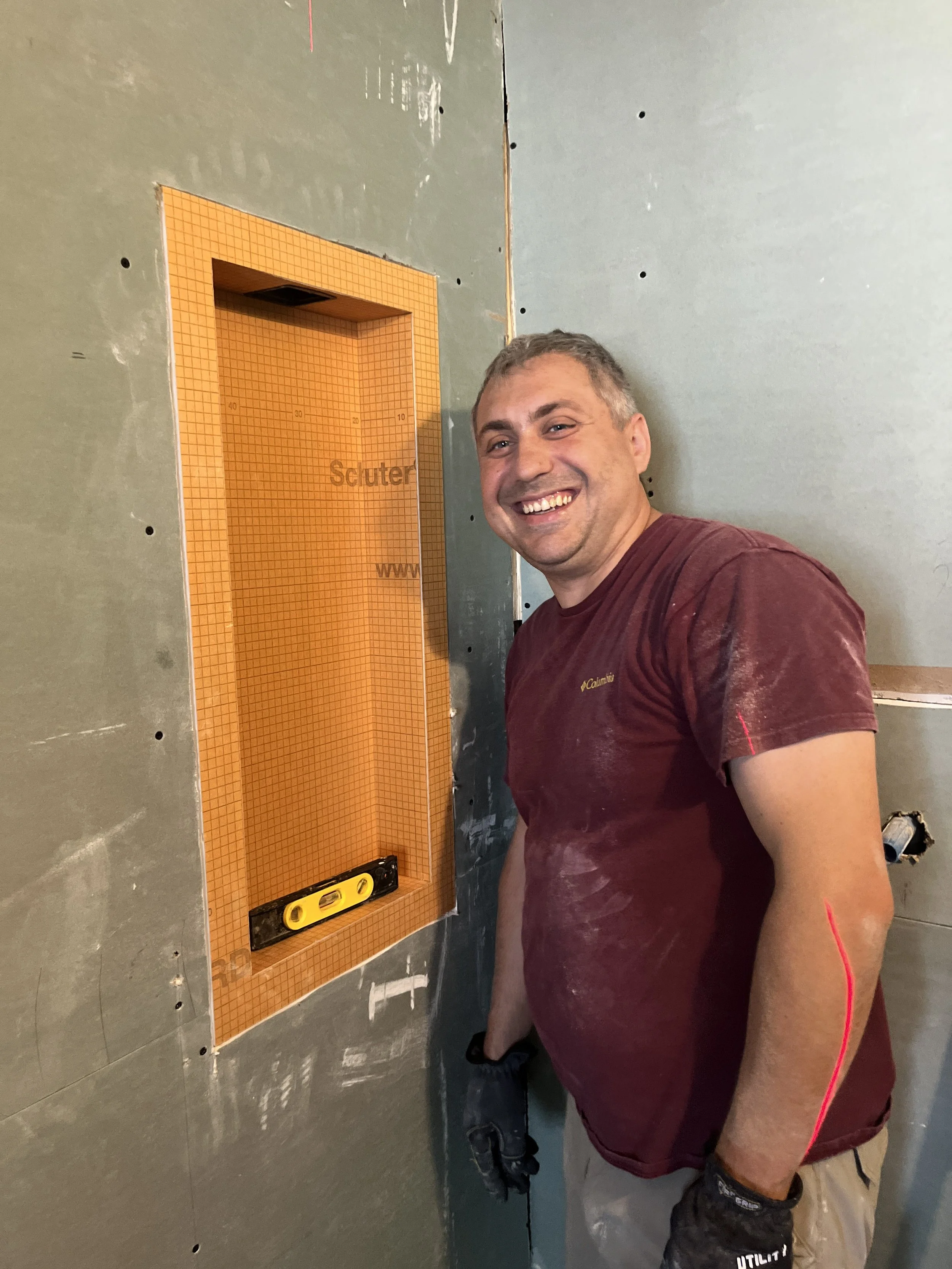 Nikolai smiling beside a newly installed recessed shower niche lined with orange Schluter waterproofing board, with a level resting on the niche shelf, during the LUX NOIR bathroom renovation by Vogman Design + Build.