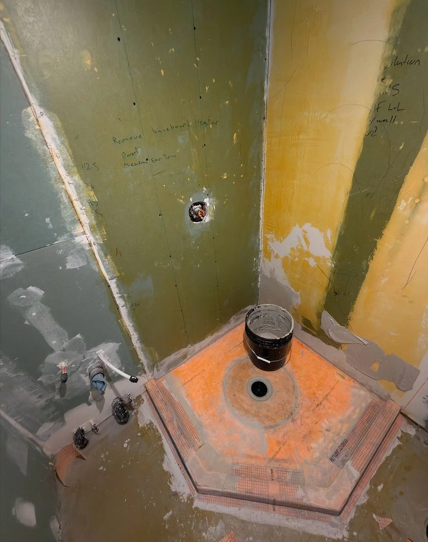 Corner shower base covered in orange waterproofing membrane with a centre drain, set against moisture-resistant drywall with the shower valve roughed in, during The Rebuild renovation by Vogman Design + Build.