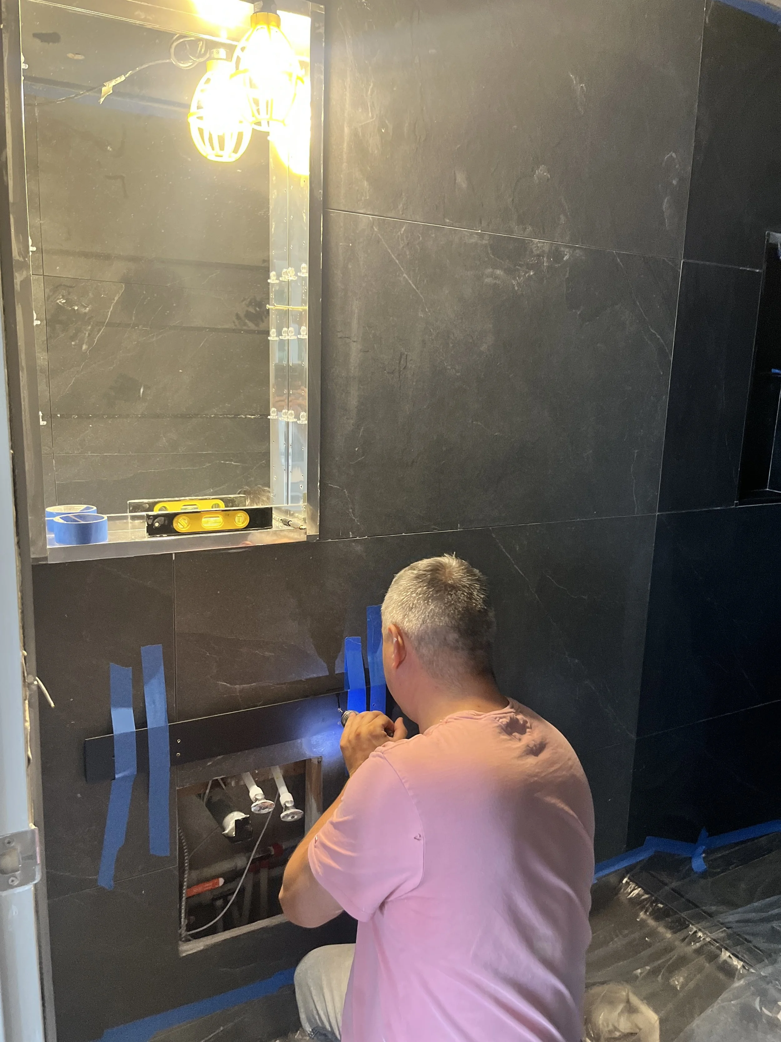 Crew member fastening a black mounting rail to the charcoal-tiled wall above the sink plumbing rough-ins, beneath a recessed mirrored cabinet with a level on its shelf, during the LUX NOIR bathroom renovation by Vogman Design + Build.