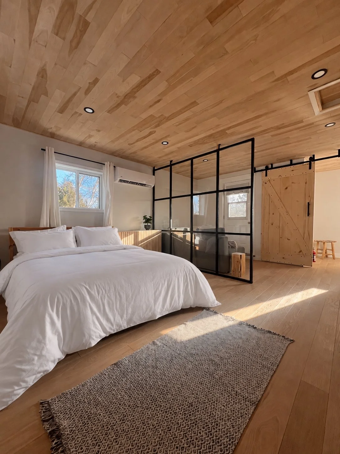 Upstairs primary bedroom in the Tobermory cottage renovation with a wood plank ceiling, white bedding, a black-framed glass partition, and a sliding barn door