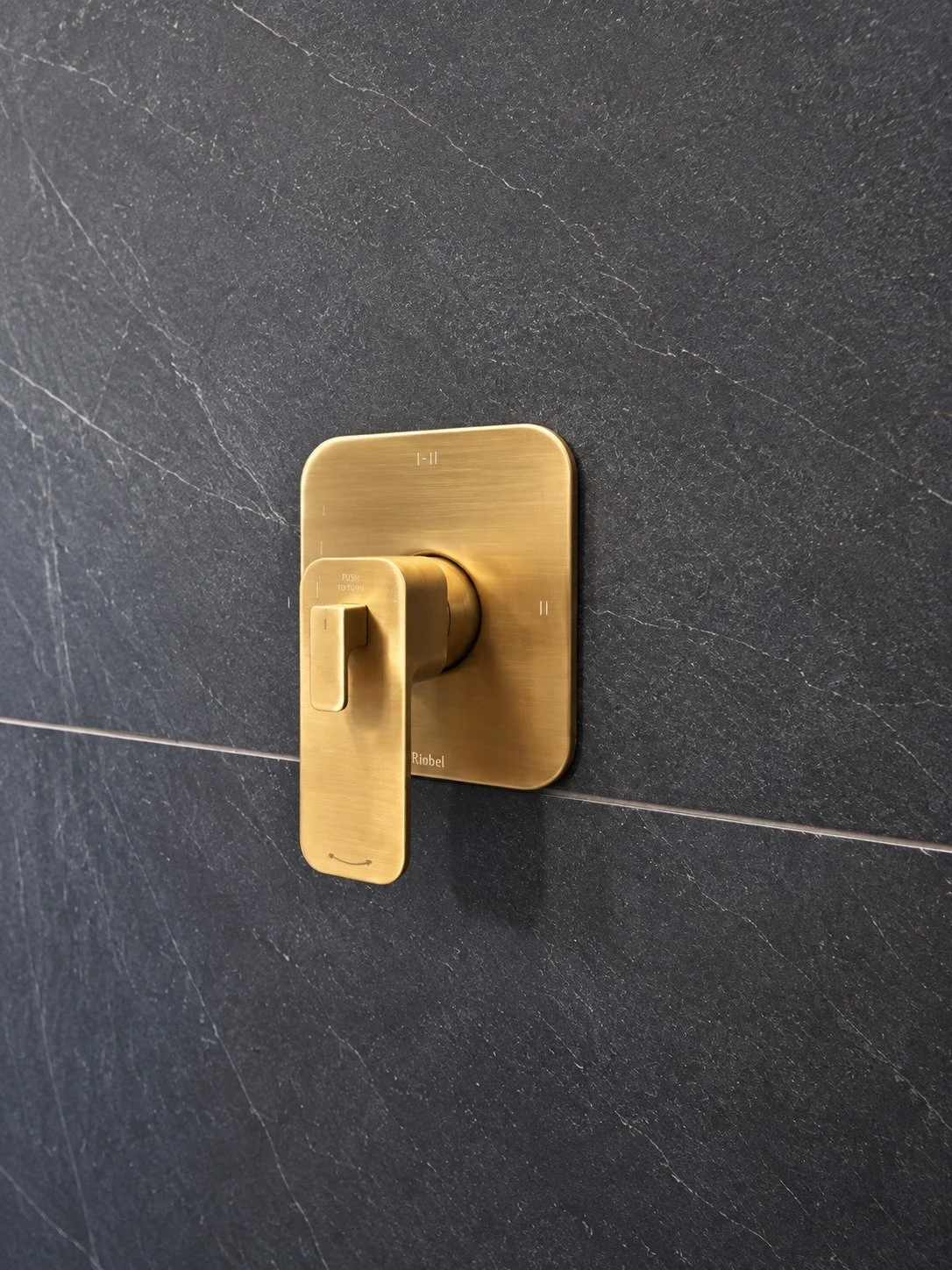 Close-up of a brushed gold Riobel shower valve with a square trim plate and lever handle, mounted on charcoal stone-look tile with white veining, in the LUX NOIR bathroom renovation by Vogman Design + Build.