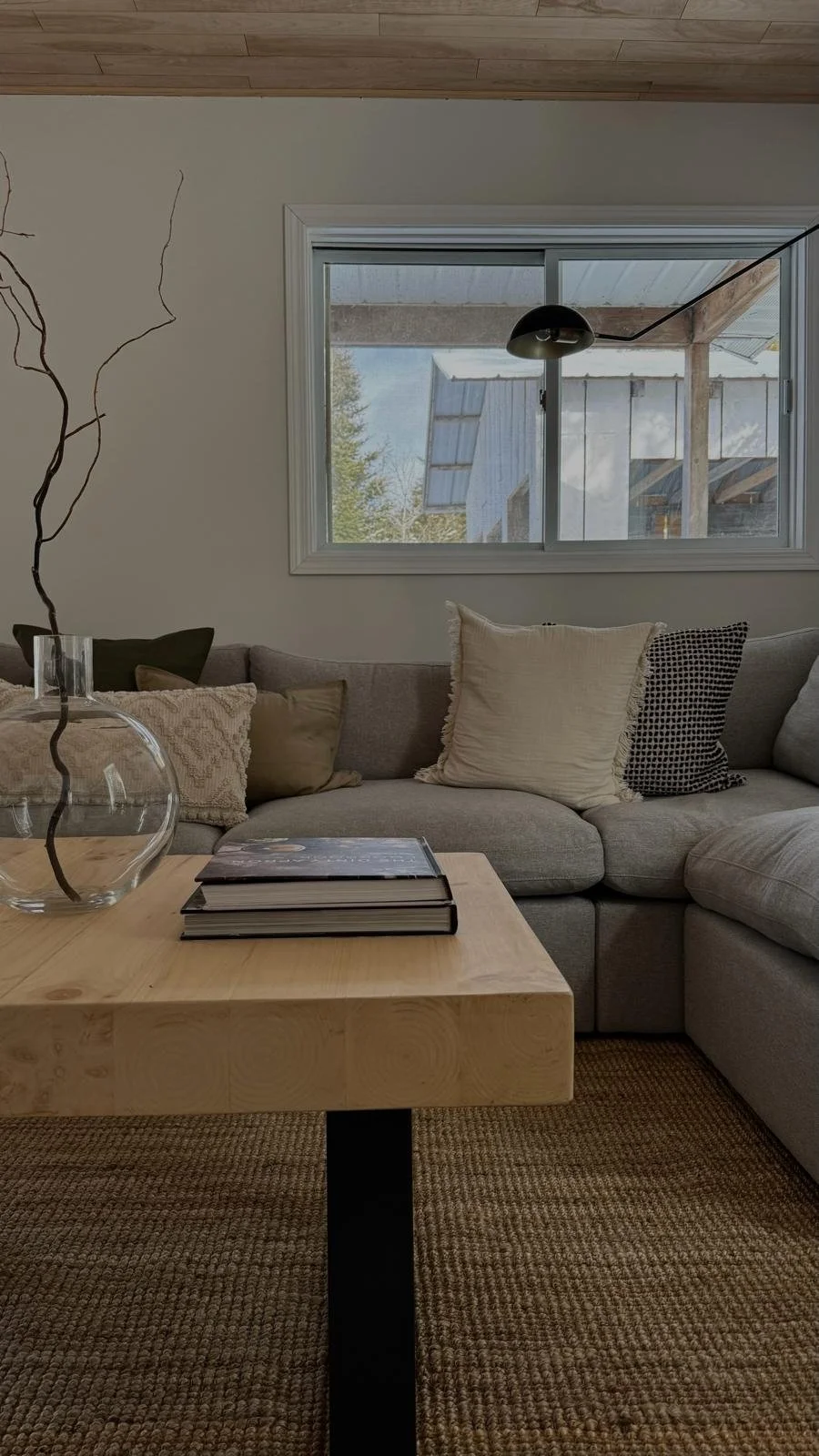 Living room in the Tobermory cottage renovation with a grey sectional sofa, live-edge wood coffee table, jute rug, and a window overlooking the property's outbuildings - Vogman Design + Build.