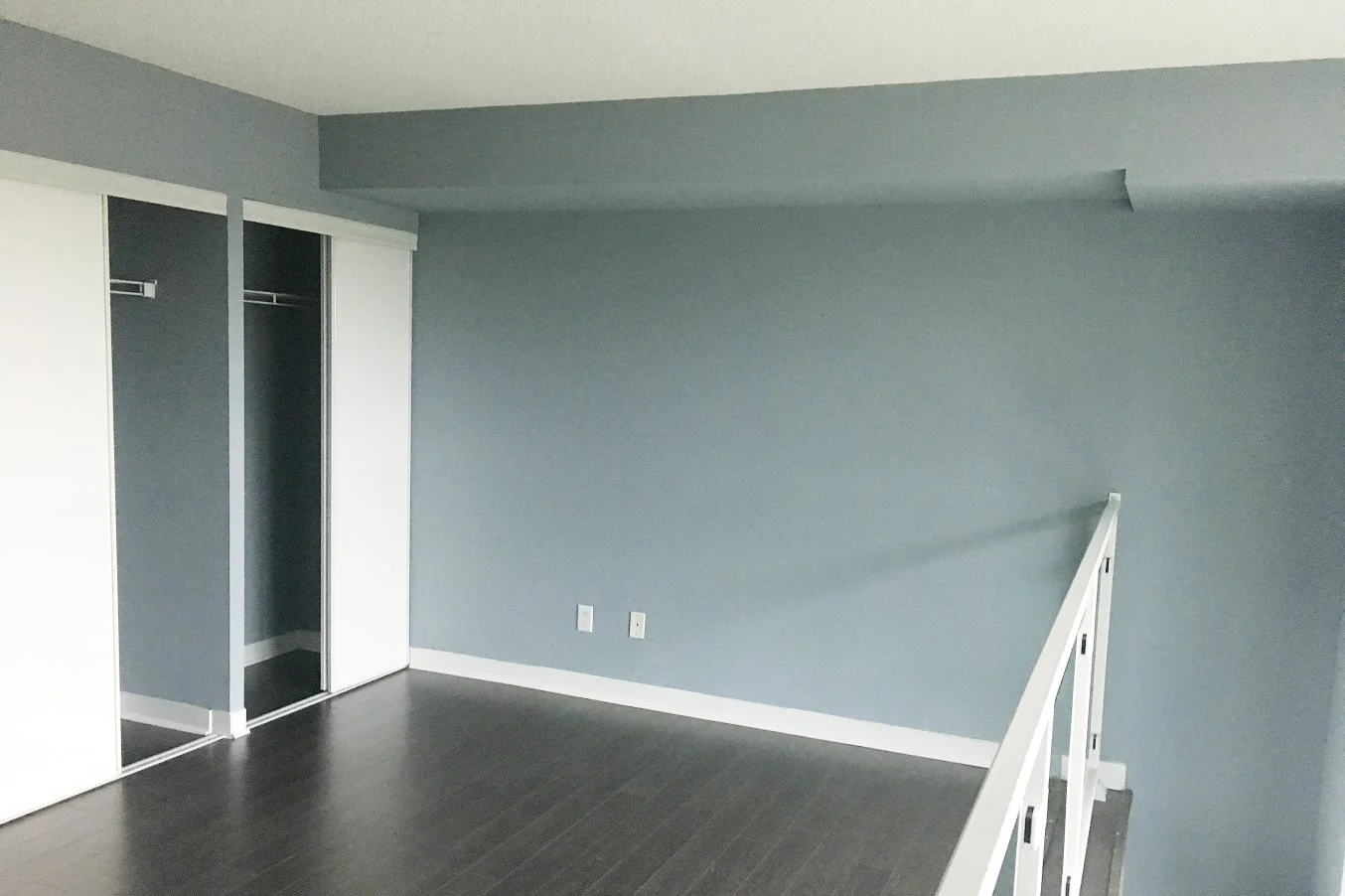 Before photo of an empty loft bedroom with blue walls, sliding closet doors, dark wood flooring, and a white glass-railed mezzanine, ahead of the full renovation