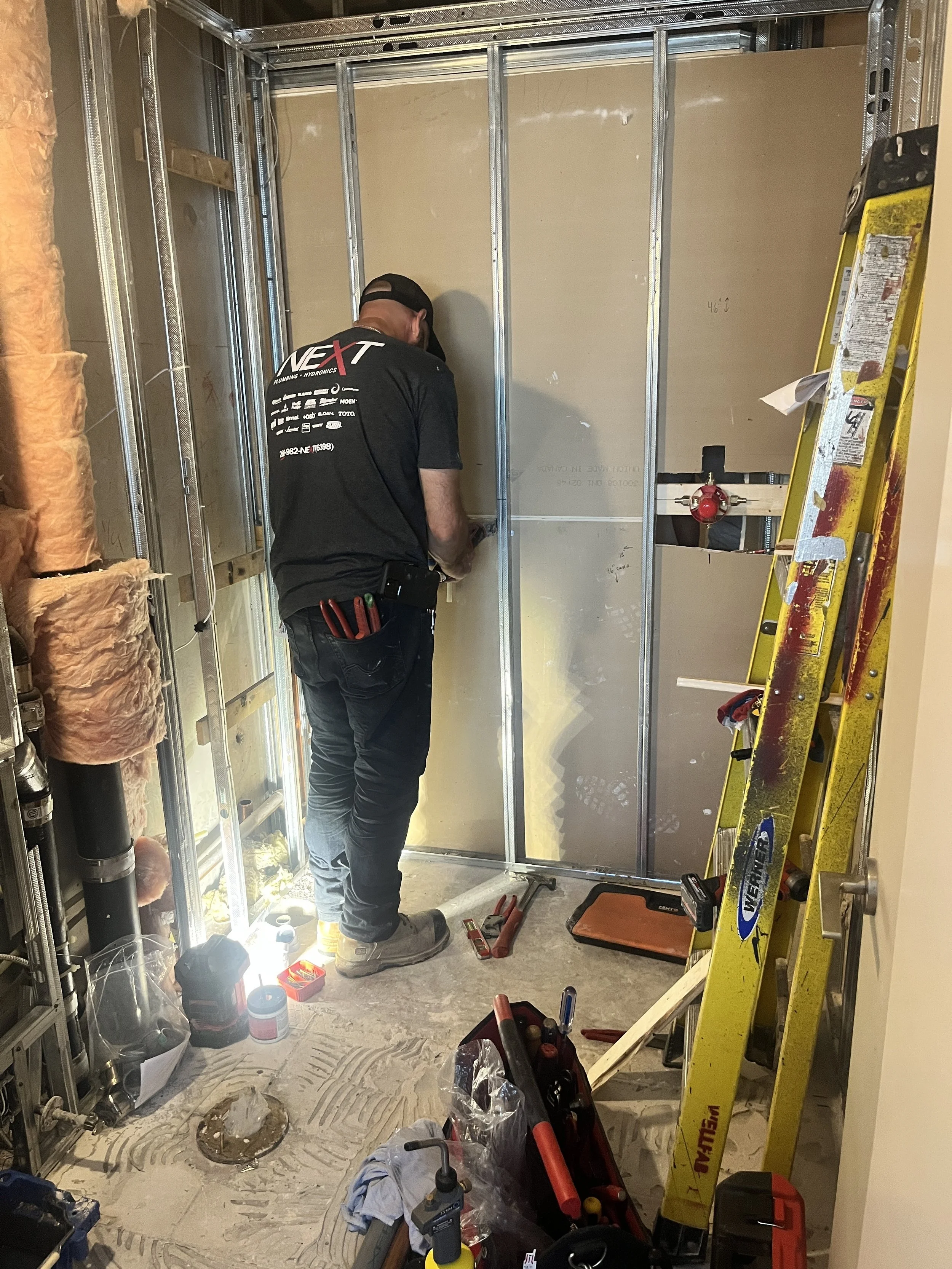 Plumber installing plumbing rough-ins inside a newly framed steel-stud shower wall, with the shower valve roughed in and tools on the floor, during the LUX NOIR bathroom renovation by Vogman Design + Build.