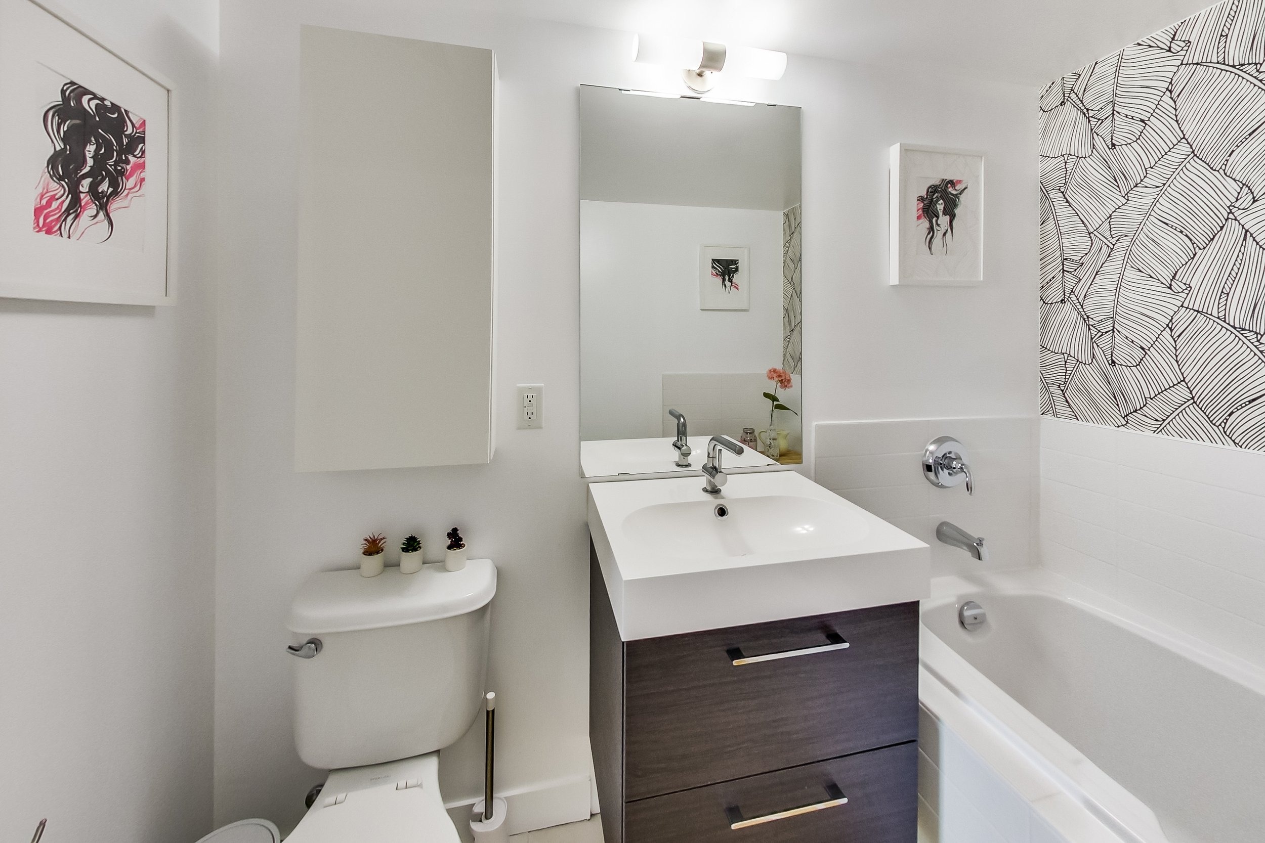Full view of the iLOFT bathroom with a dark wood vanity, white countertop sink, framed artwork, and black and white leaf-print wallpaper above the tub